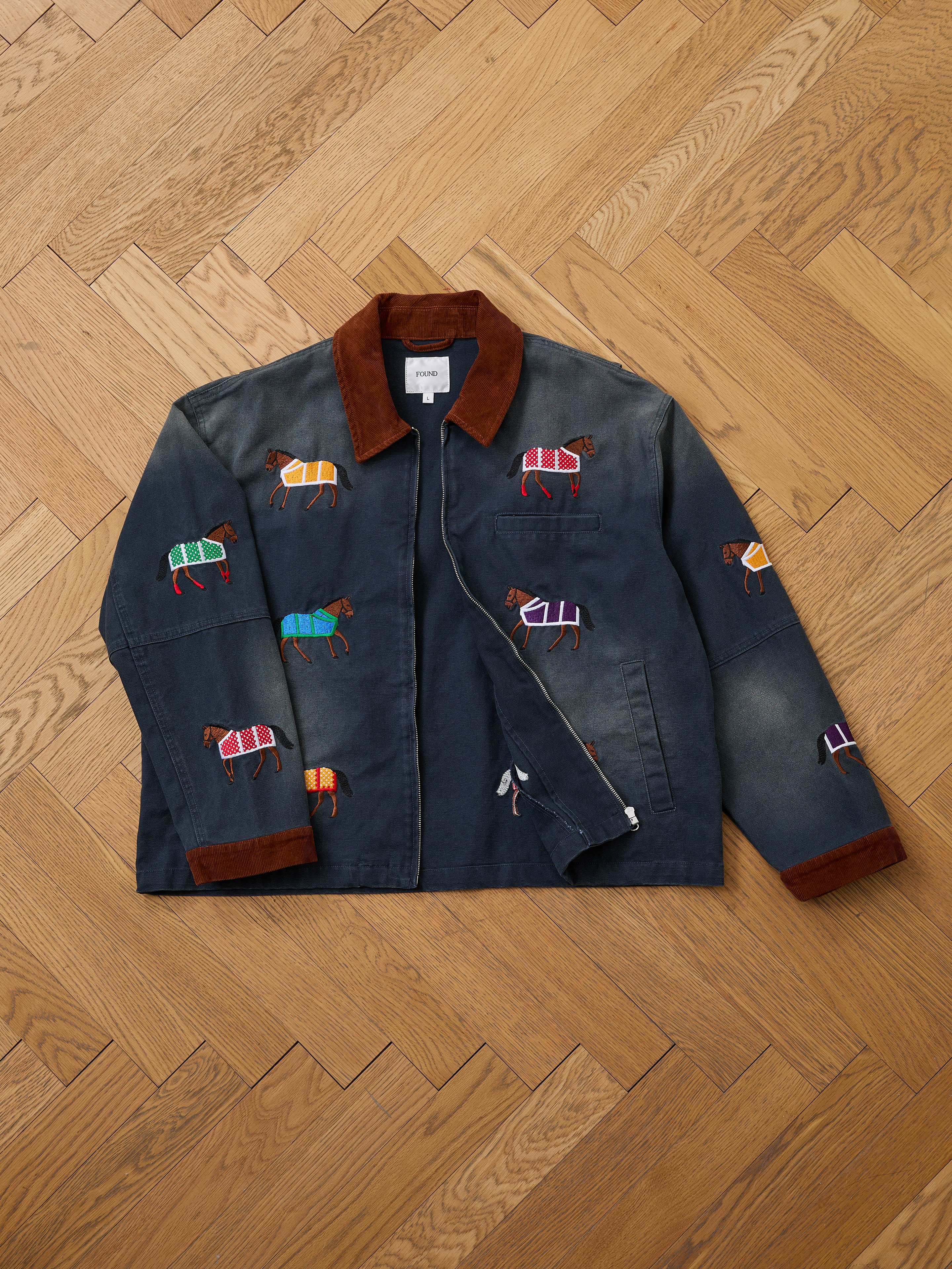 The Found Lariat Horse Embroidered Work Jacket, a unisex corduroy piece with brown collar and cuffs and colorful horse embroidery, is displayed flat on a wooden herringbone floor.