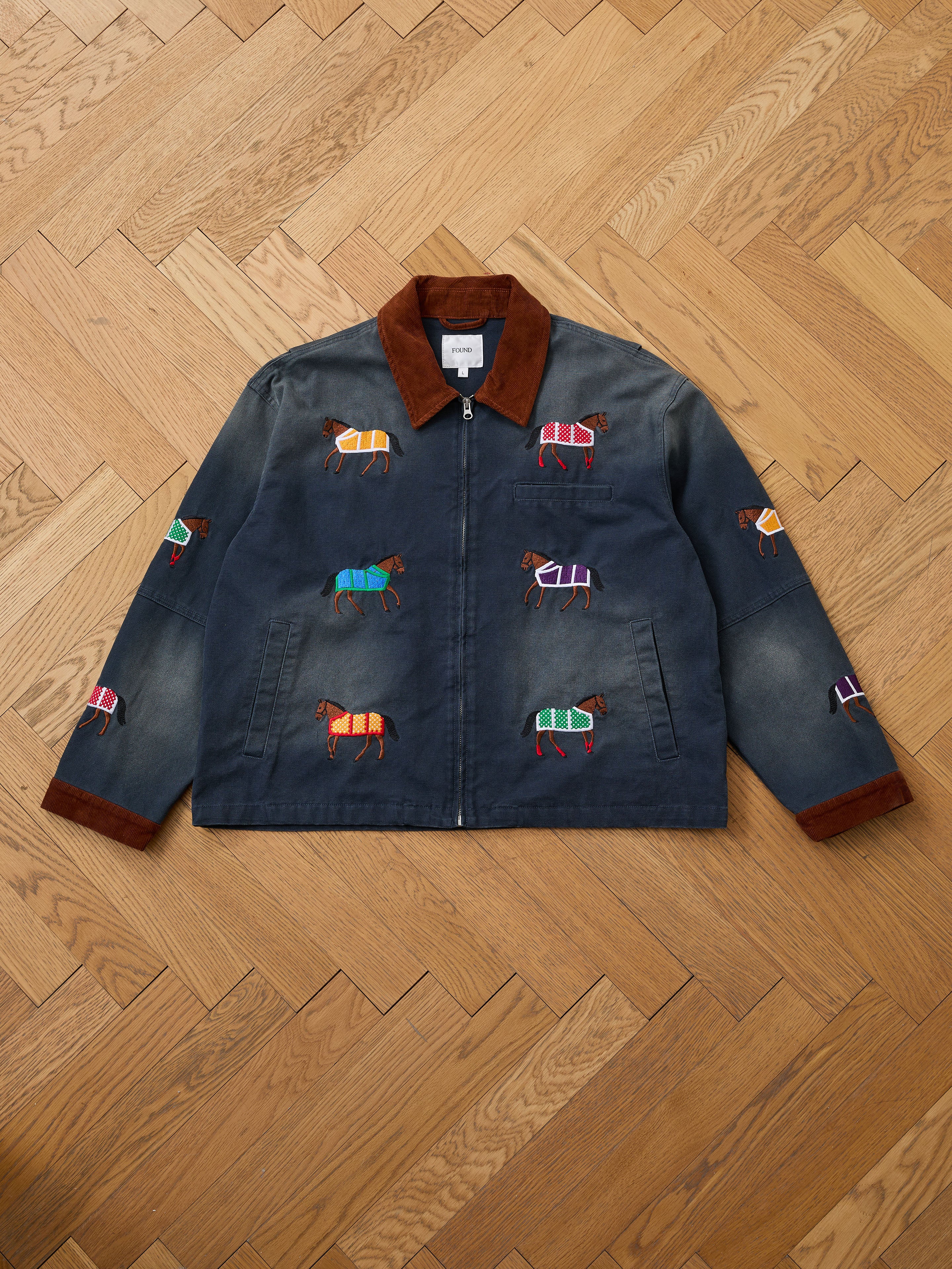 The Found Lariat Horse Embroidered Work Jacket, featuring dark denim, a brown collar, zip front, and colorful horse embroidery, is displayed on a wooden herringbone floor.