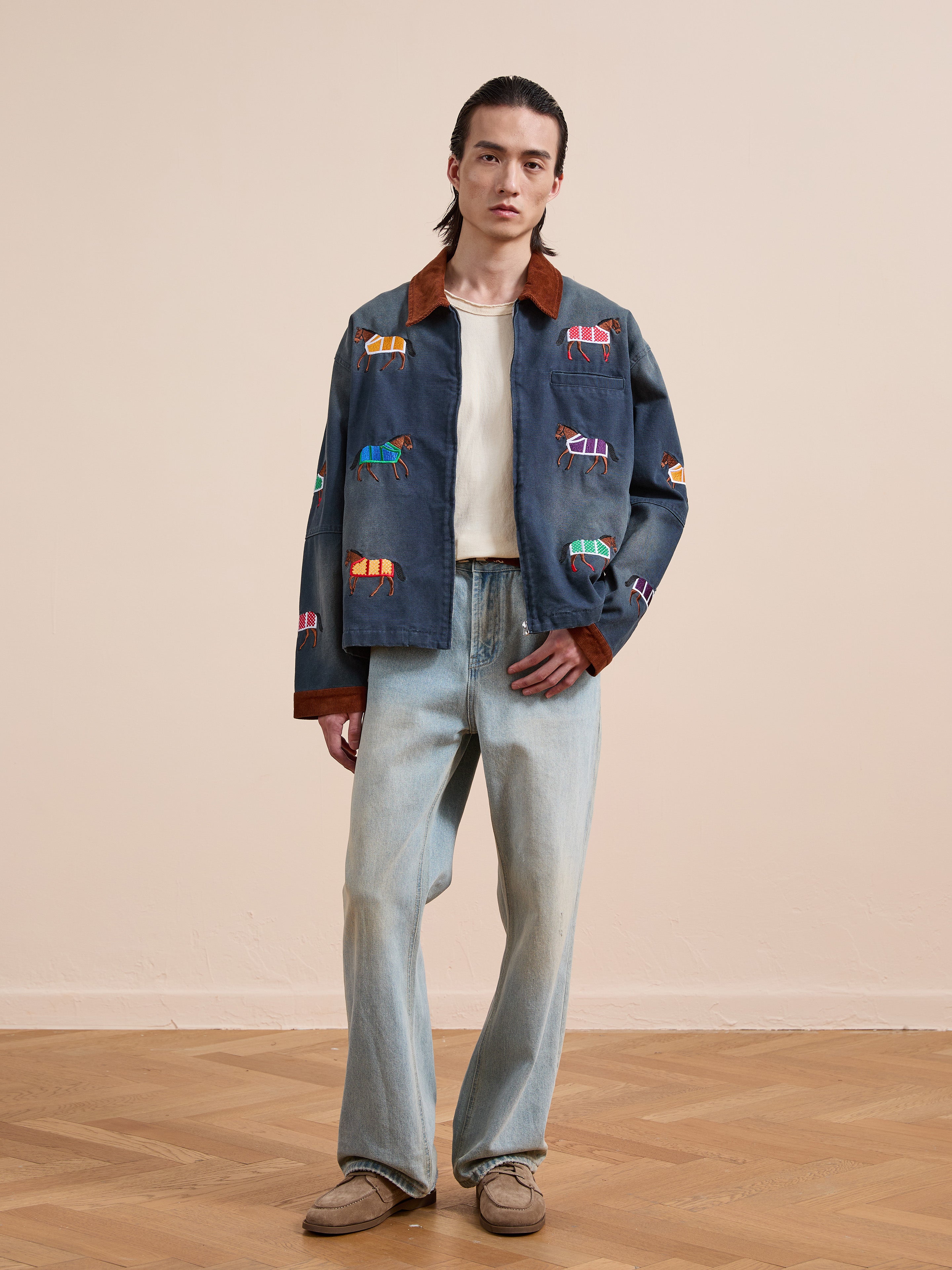 A person stands indoors on a wooden floor, wearing light blue jeans, a white shirt, tan shoes, and the Found Lariat Horse Embroidered Work Jacket.