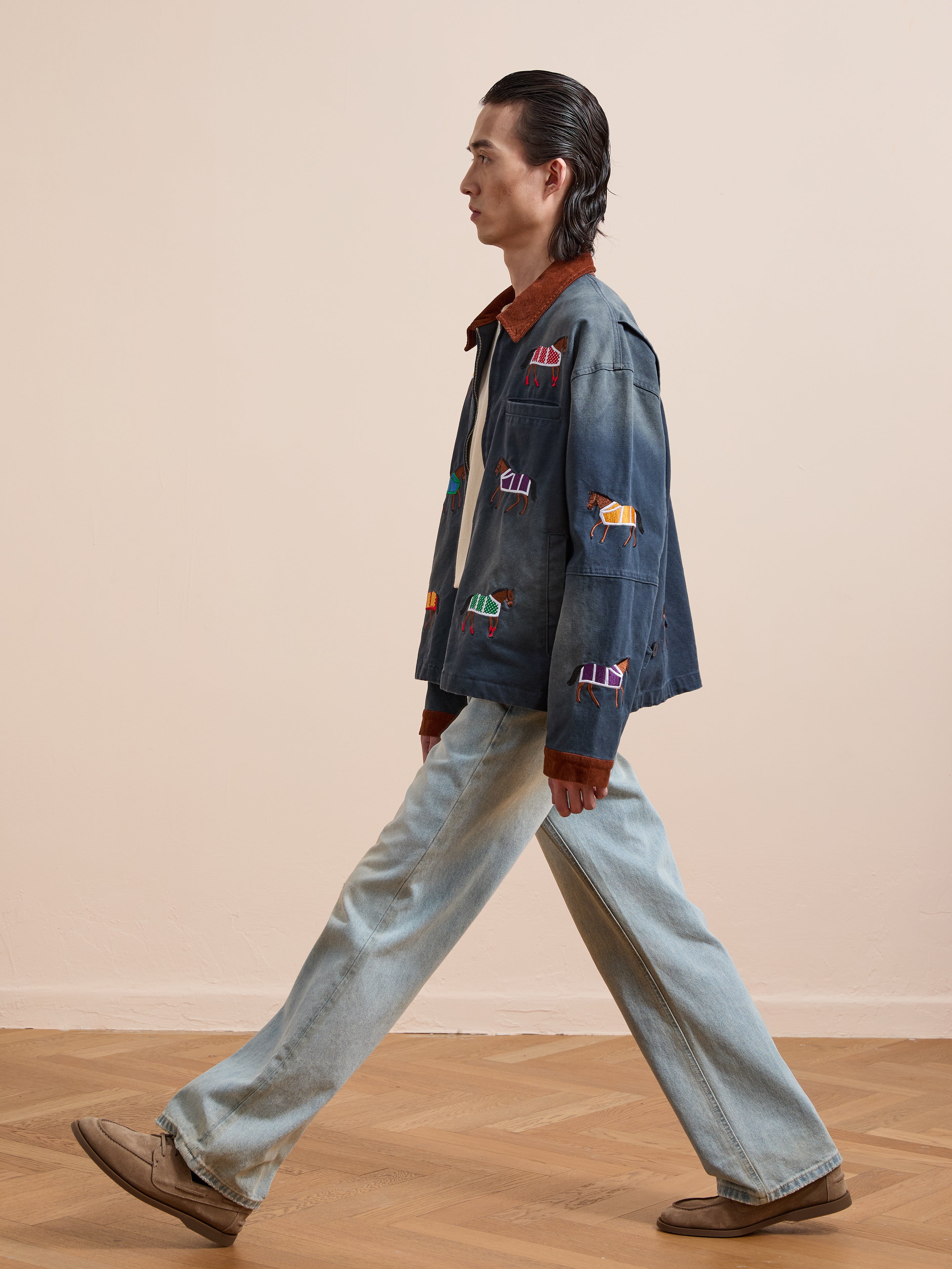 A person with long dark hair wears the Found Lariat Horse Embroidered Work Jacket in navy, wide-leg jeans, and beige shoes while walking indoors on a wooden floor.