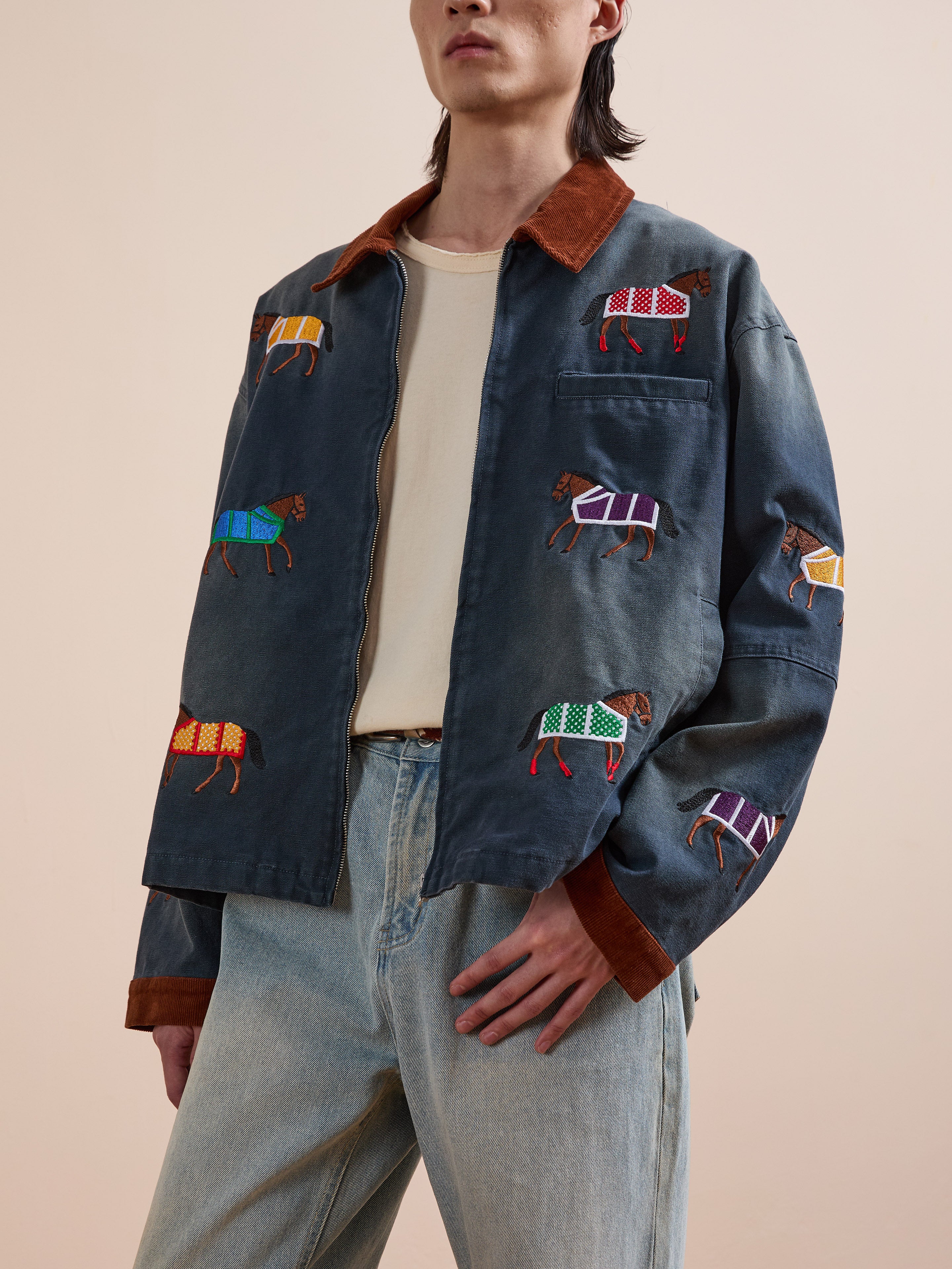 A person wears the Found Lariat Horse Embroidered Work Jacket, featuring vibrant horse embroidery, paired with a beige shirt and light blue jeans, standing against a plain background.