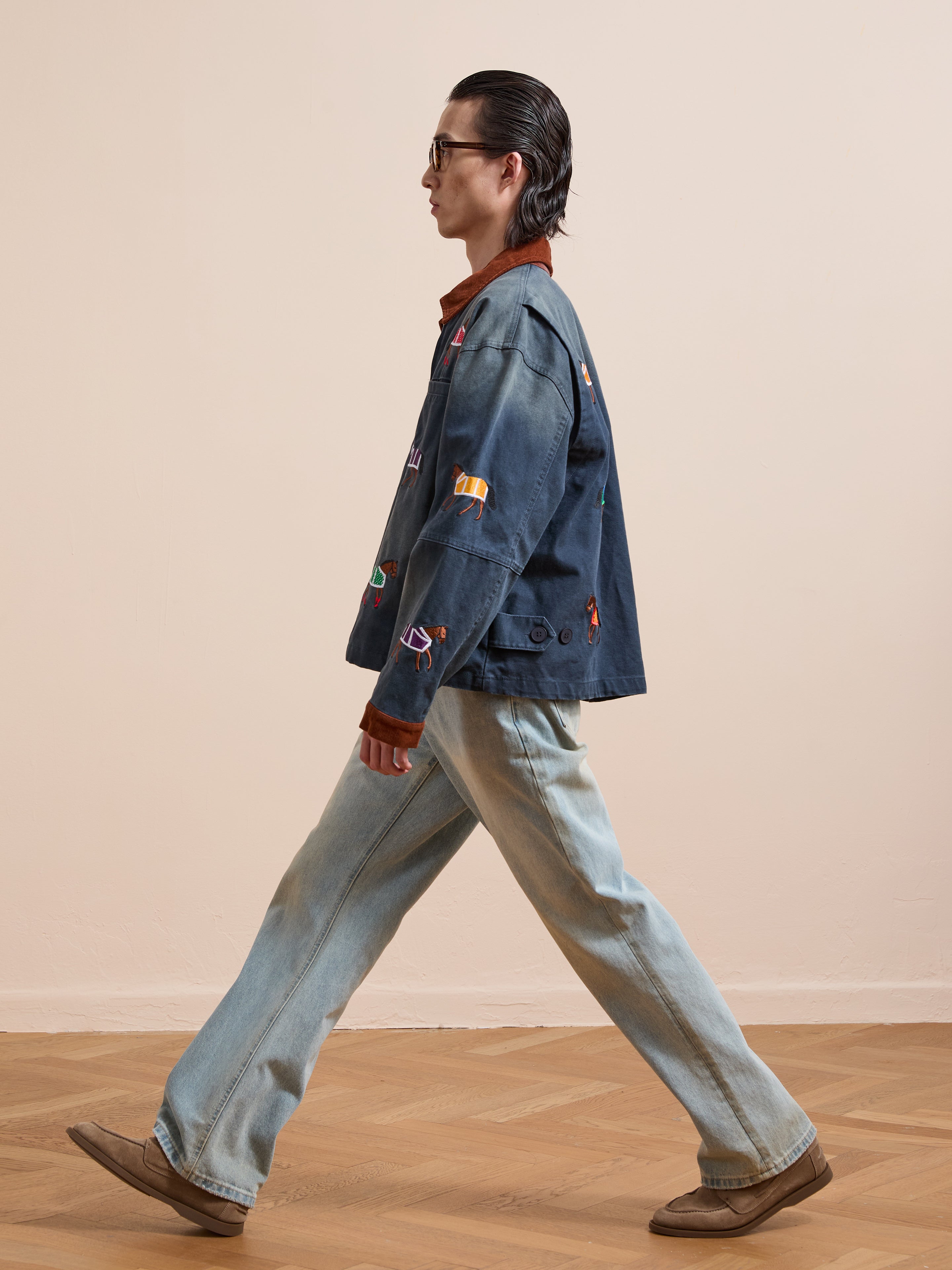 Wearing Found’s Lariat Horse Embroidered Work Jacket with colorful patches, light jeans, beige shoes, and sunglasses, a person walks past a plain beige wall on a wooden floor.
