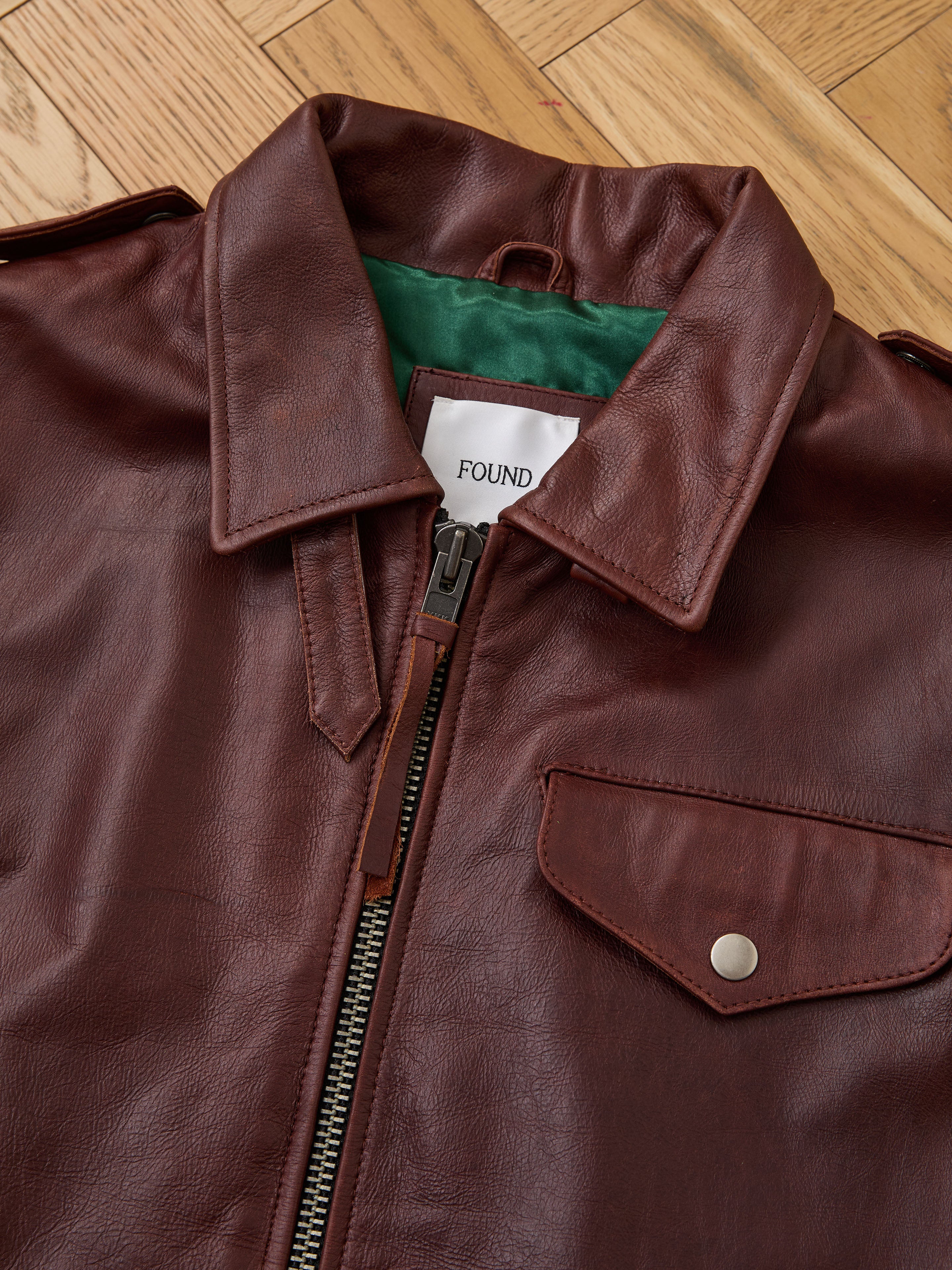 The Found Leather Pocket Vest, featuring a distressed brown finish, zipper front, utility pockets with a flap and snap button, and green inner lining, is laid flat on a wooden floor.