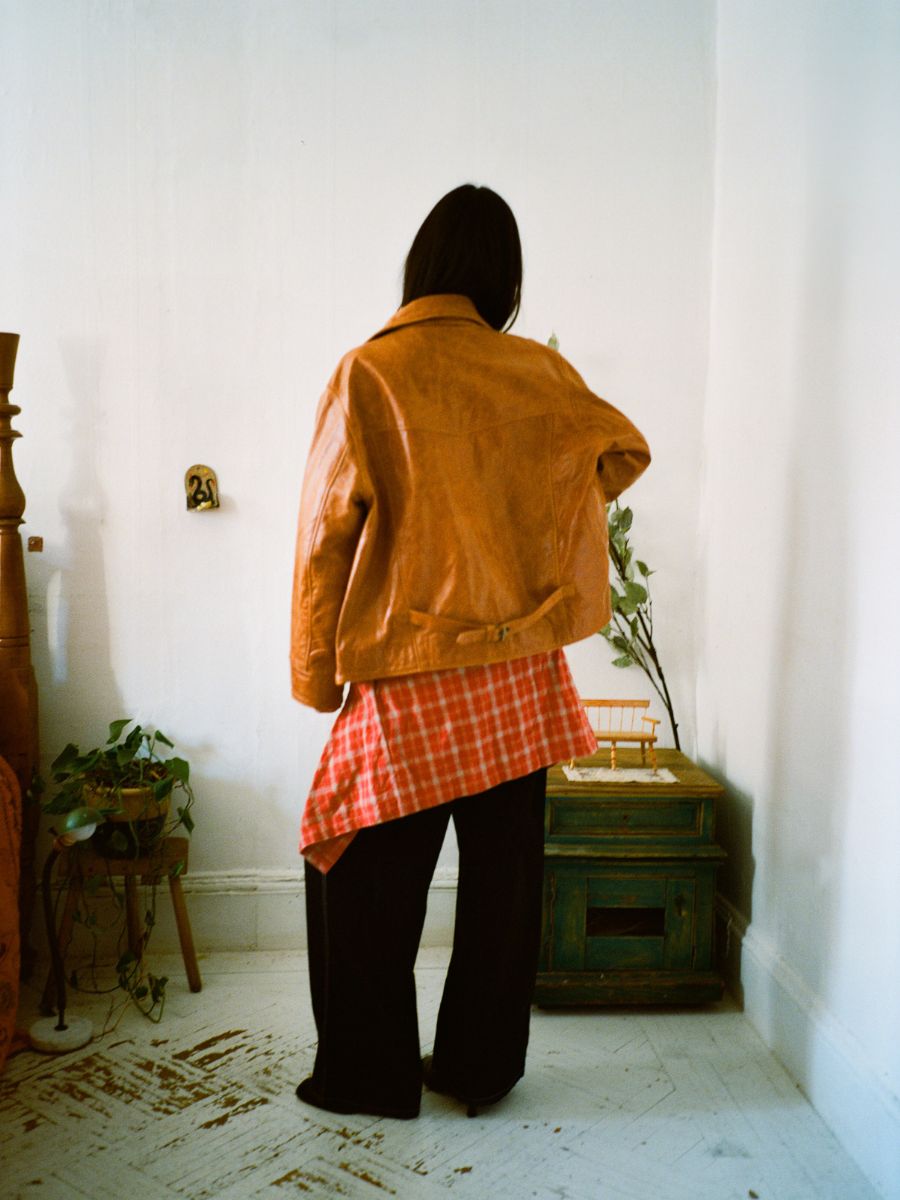 A person faces away from the camera, wearing a Marlboro Sun Faded Leather Racer Jacket by FOUND over a red plaid shirt. The white walls and lush plants in the room enhance the rugged appeal of this unisex leather piece.
