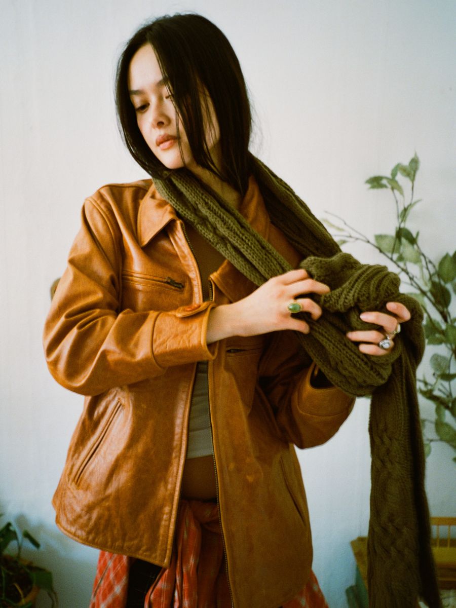 A person in a Marlboro Sun Faded Leather Racer Jacket by FOUND adjusts a green scarf with a white wall and plant in the background.