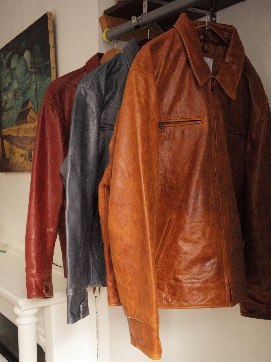 A rack showcases three leather jackets in brown, blue, and red, with the FOUND Marlboro Sun Faded Leather Racer Jacket standing out. A framed painting on the wall behind adds elegance to the display.