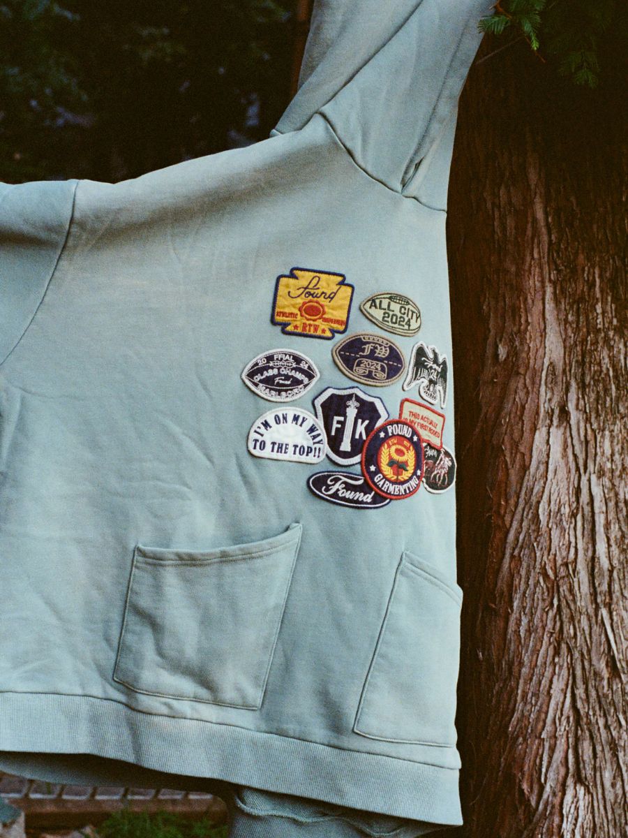 The FOUND Multi Patch Hoodie, made from vintage French Terry Cotton and embellished with vibrant patches, hangs gracefully over a tree branch.