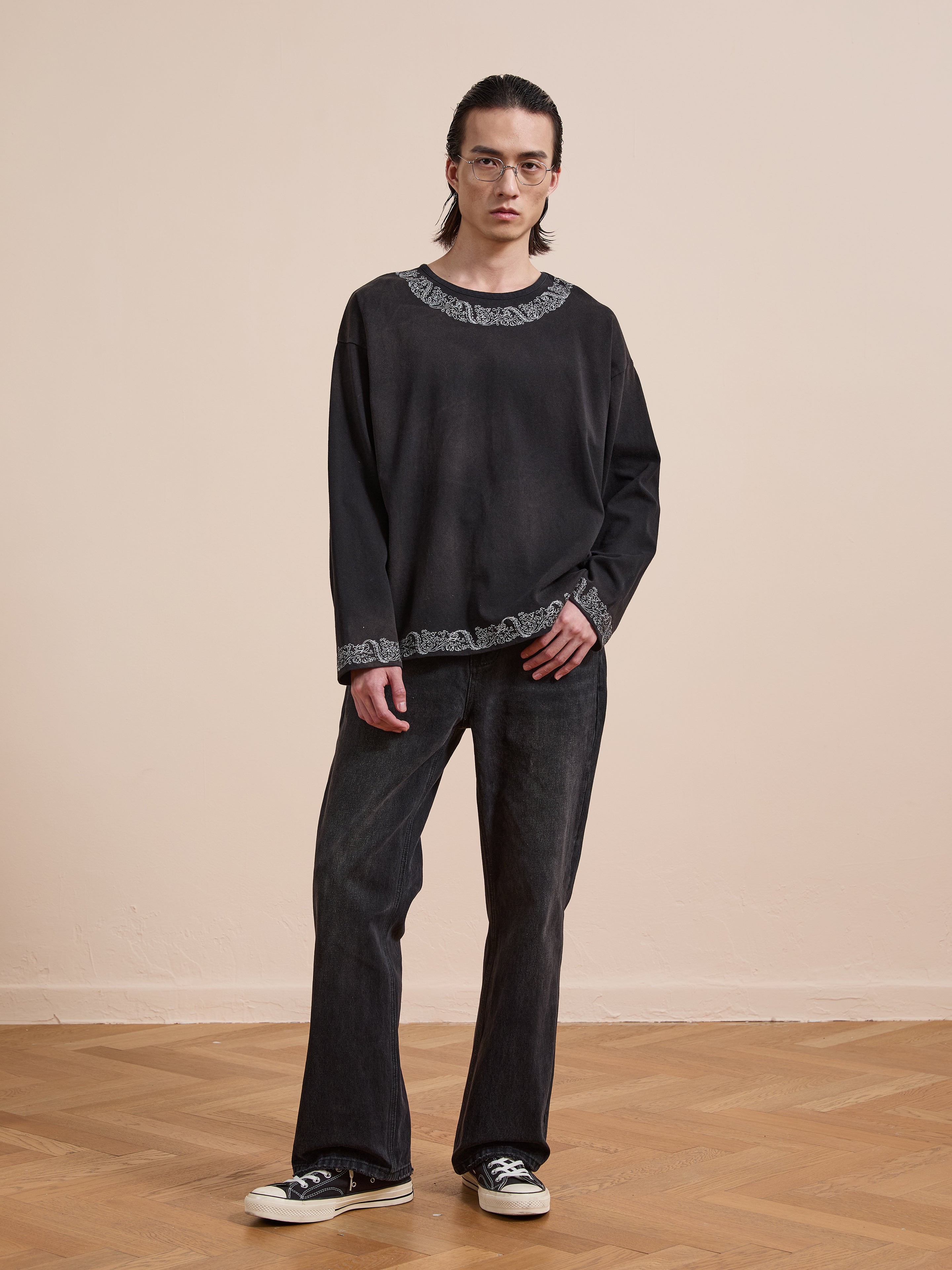 A person with long dark hair, glasses, and a neutral expression stands indoors wearing the FOUND Metallic Long Sleeve Embroidered Tee, styled with black jeans and black sneakers.
