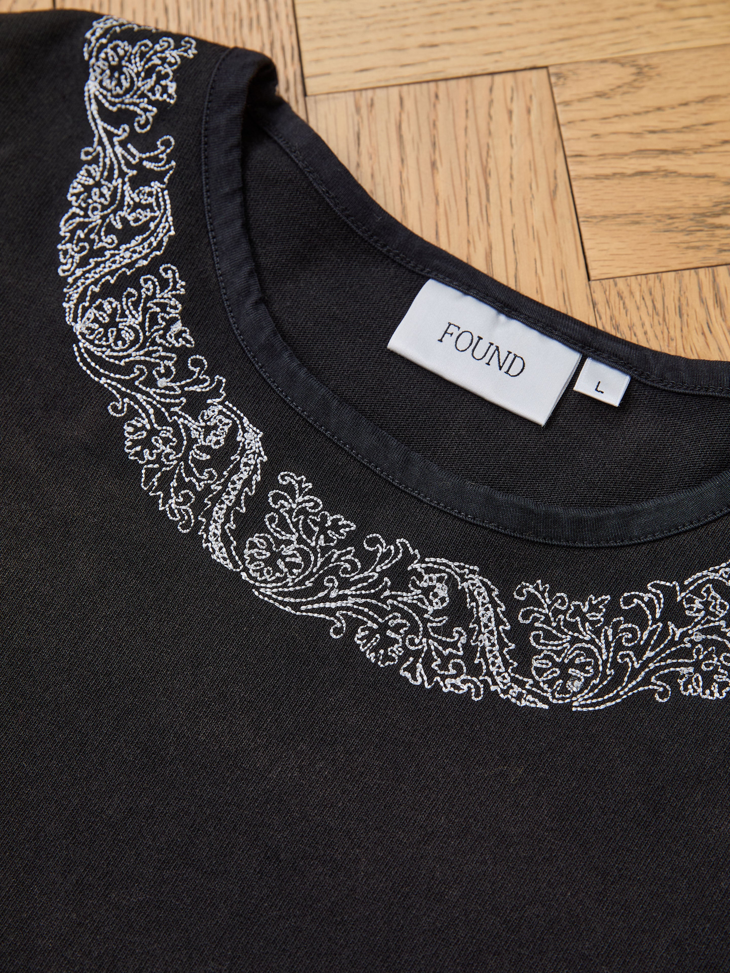 A FOUND Metallic Long Sleeve Embroidered Tee in size L, featuring white embroidery along the neckline and a relaxed unisex fit, is displayed flat on a wooden surface.