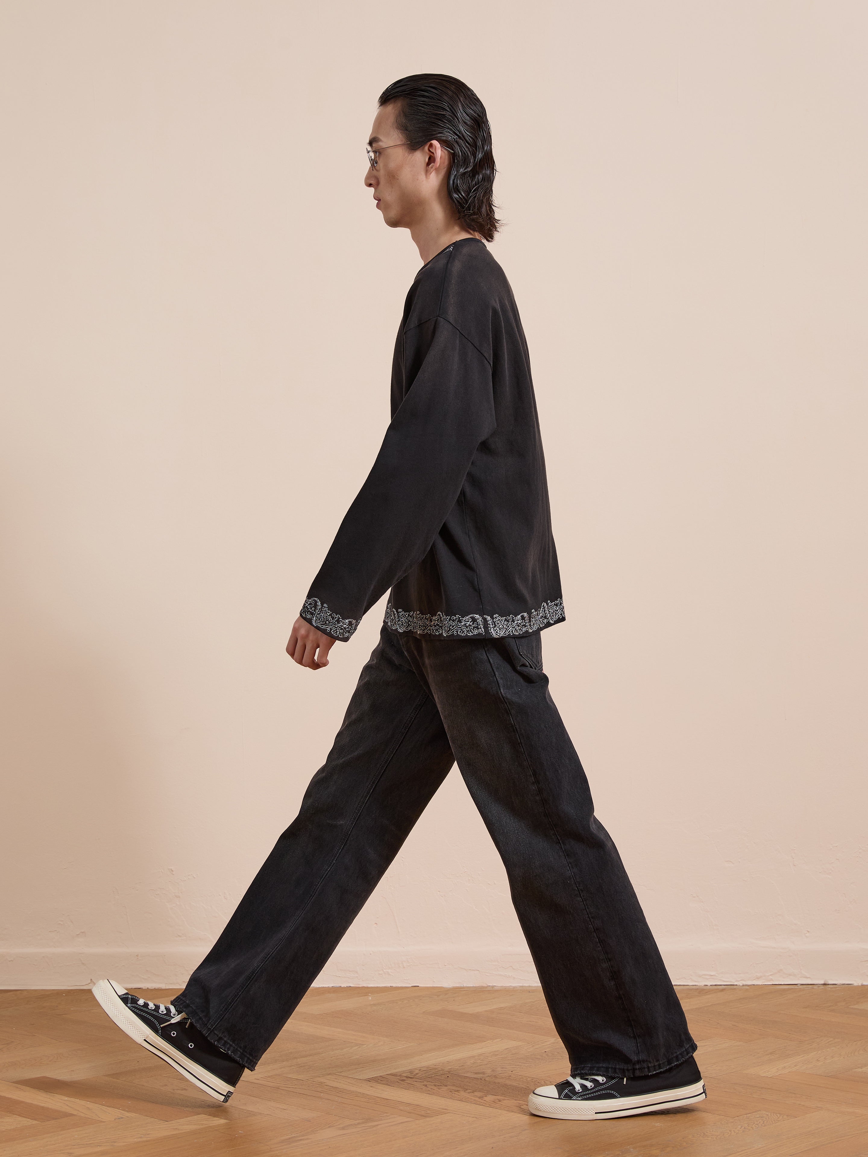 A person with long dark hair and glasses wears the FOUND Metallic Long Sleeve Embroidered Tee, black wide-leg jeans, and black sneakers while walking indoors on a wooden floor.
