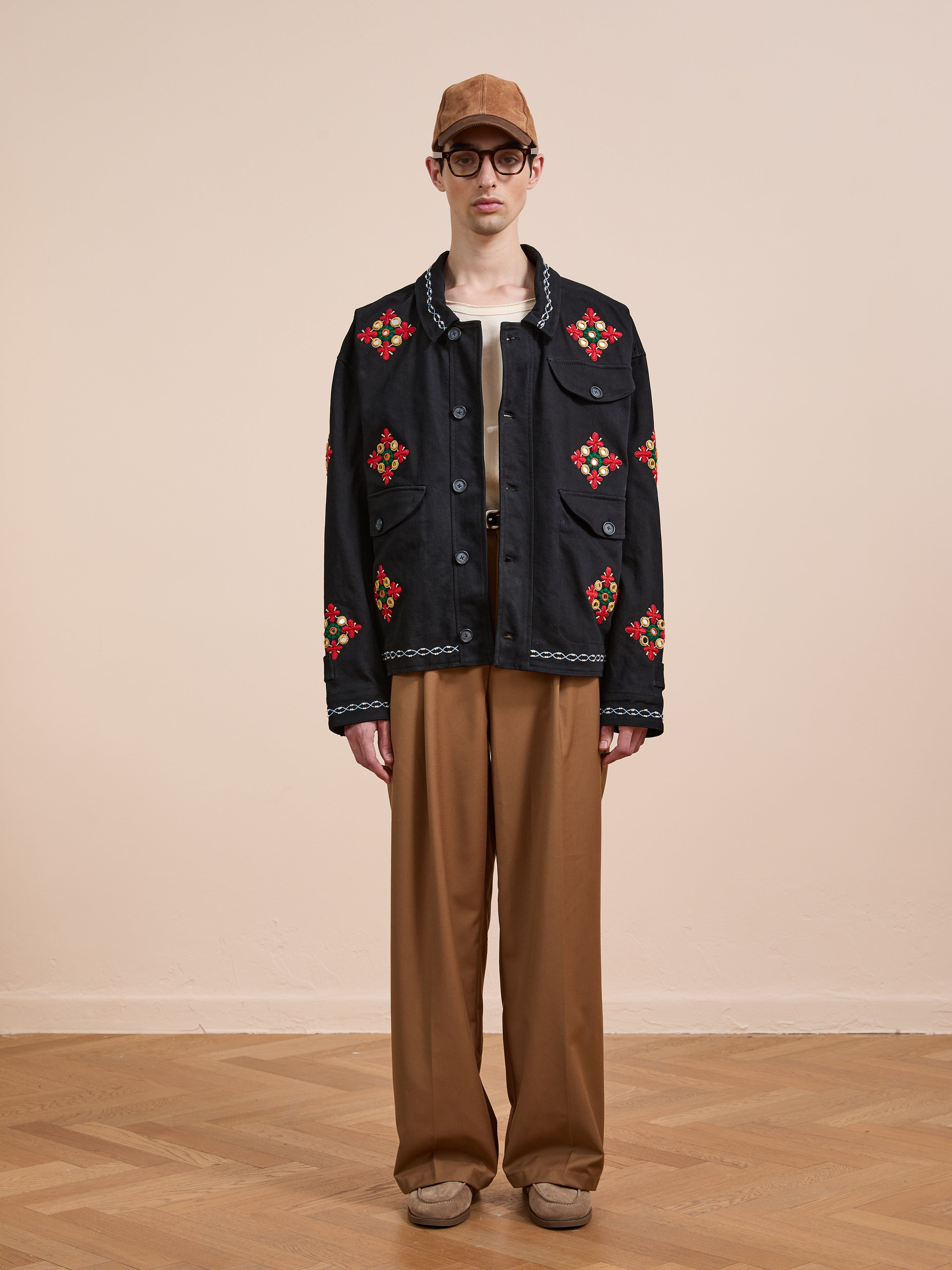 Indoors, a person wears a brown cap, glasses, beige shirt, brown wide-leg pants, beige shoes, and the Found Mirror Ornament Hand Embroidered Work Jacket—a black cotton piece with red and green embroidery—against a plain background.
