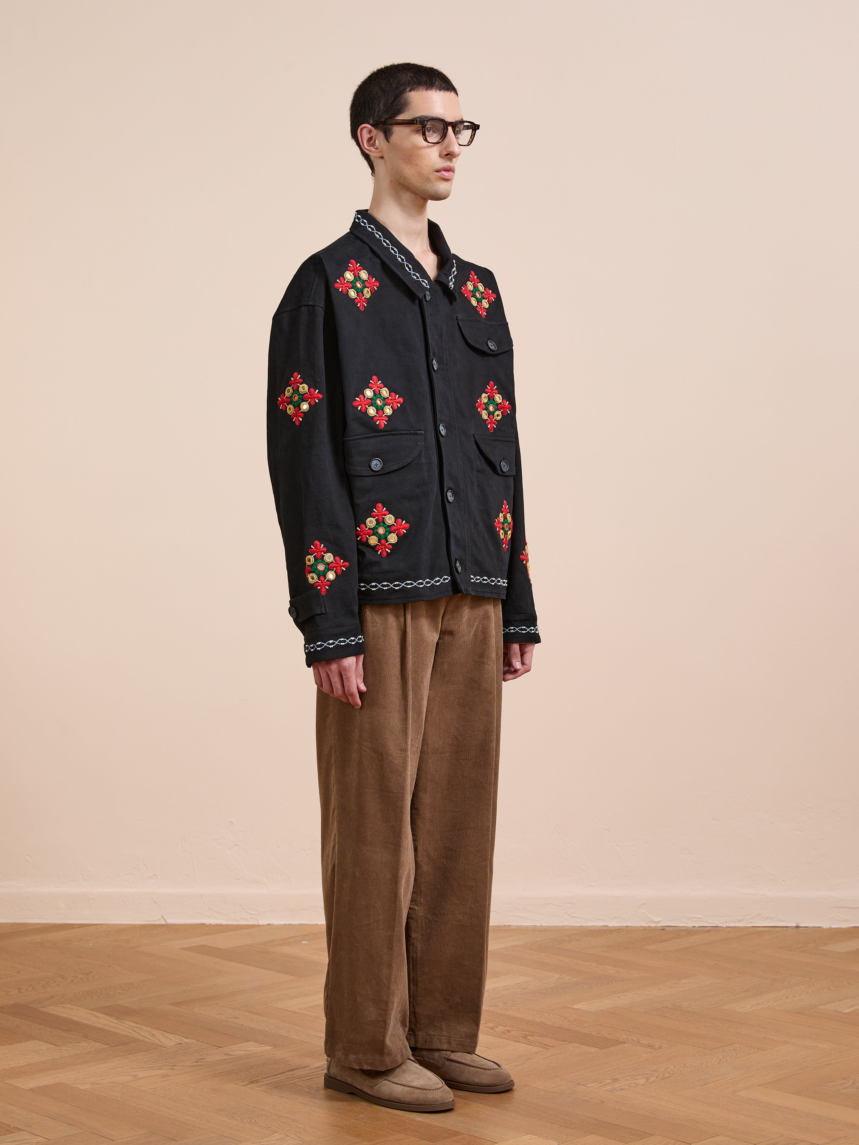 A person wearing glasses, Found's Mirror Ornament Hand Embroidered Work Jacket, brown pants, and beige shoes stands indoors on a wooden floor against a plain beige wall.