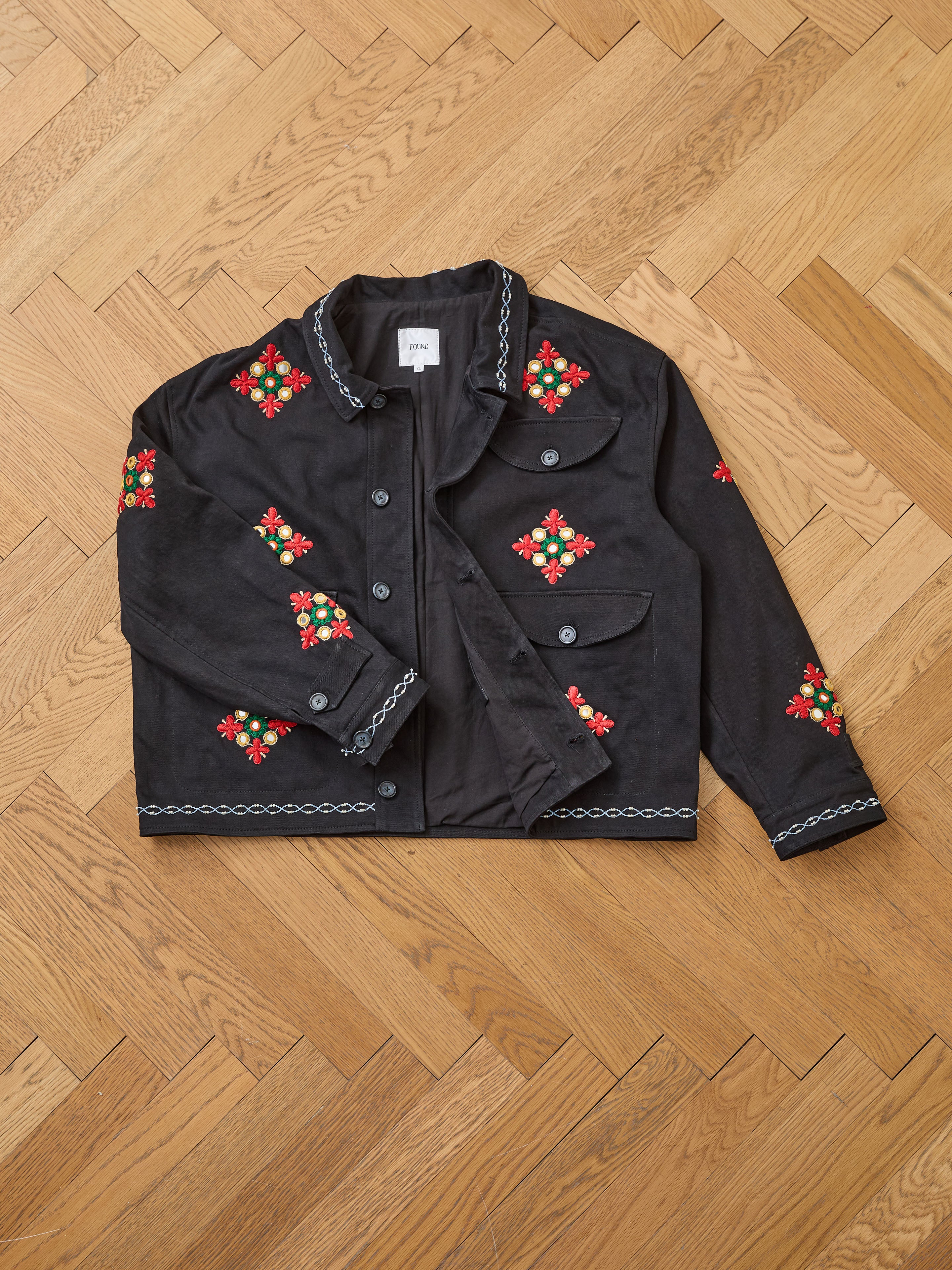 The Found Mirror Ornament Hand Embroidered Work Jacket, a handcrafted black cotton piece with red, green, and white floral embroidery on the front, sleeves, and collar, is displayed on a wooden herringbone floor.