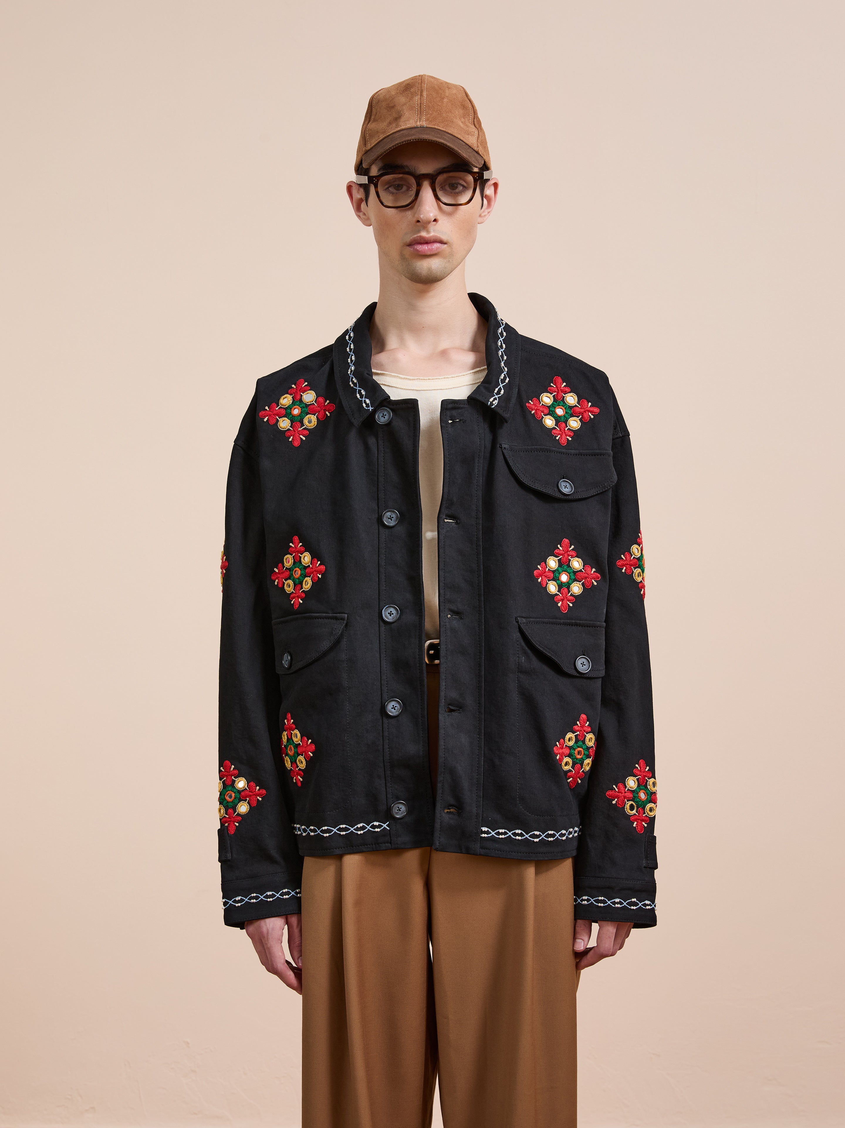 A person wearing a brown cap, glasses, beige shirt, brown pants, and the Found Mirror Ornament Hand Embroidered Work Jacket with black fabric and red-green floral embroidery stands against a plain light background.