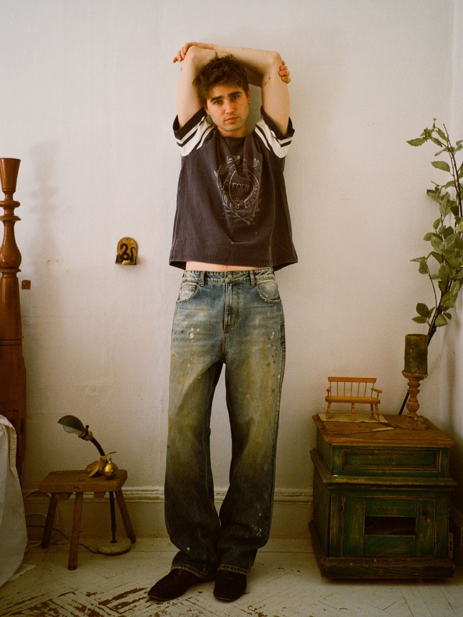 Dressed in FOUND's Monet Paint Jeans for a creative flair, a person stands indoors with their arms stretched above head. The setting is minimalist, highlighted by a lush plant and a small green cabinet.