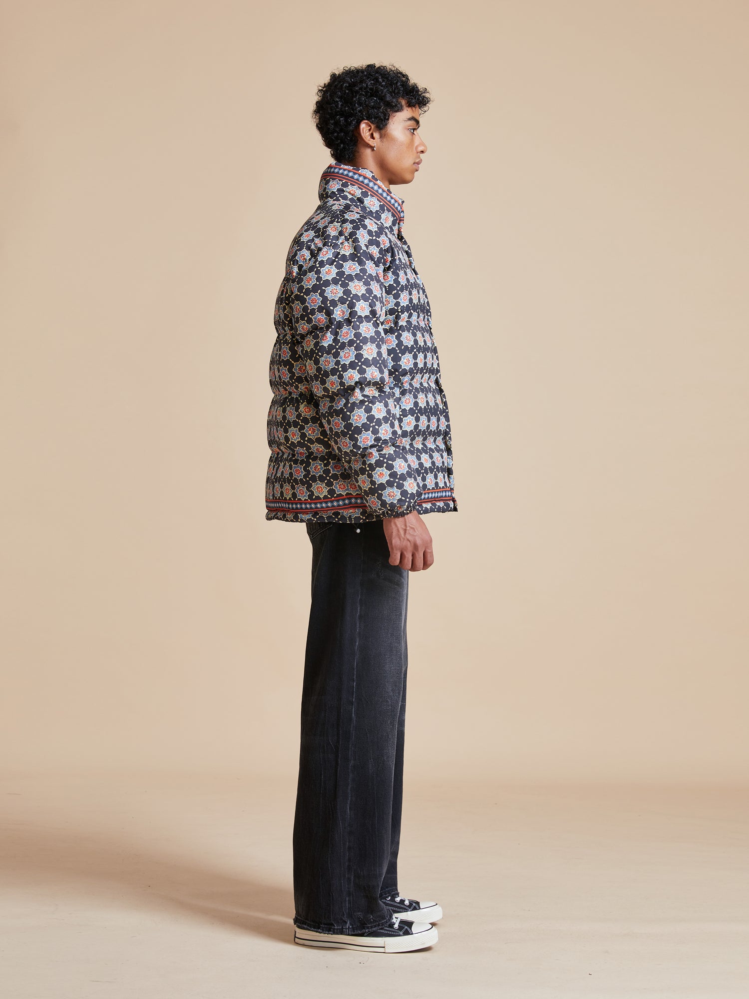 A man in a black jacket and jeans standing in front of a wall, wearing a Found Ajrak Block Puffer Jacket showcasing Pakistani block-printed patterns.