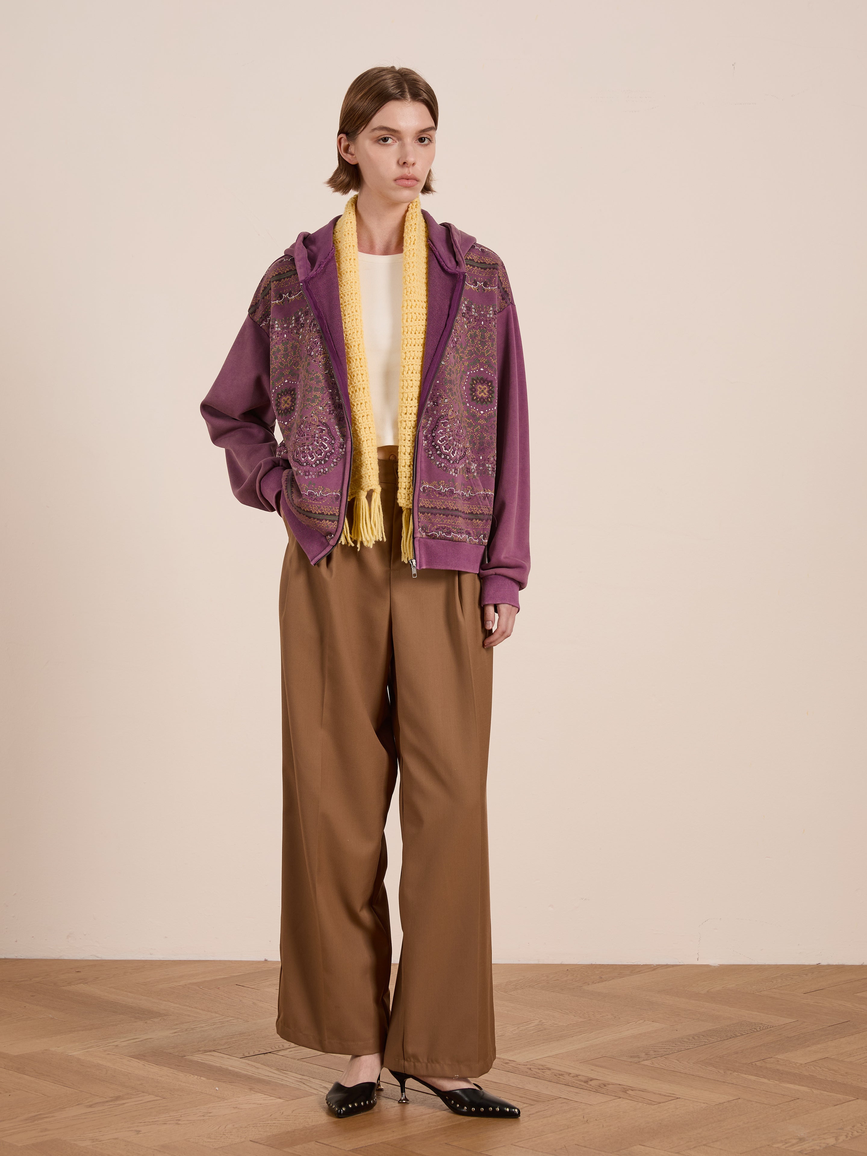 Dressed in a purple Motif Zip-Up Hoodie by FOUND, reminiscent of mandala art, with a yellow scarf, cream top, and brown wide-leg pants, an individual stands against a plain beige wall on a parquet floor, effortlessly embodying streetwear essentials.