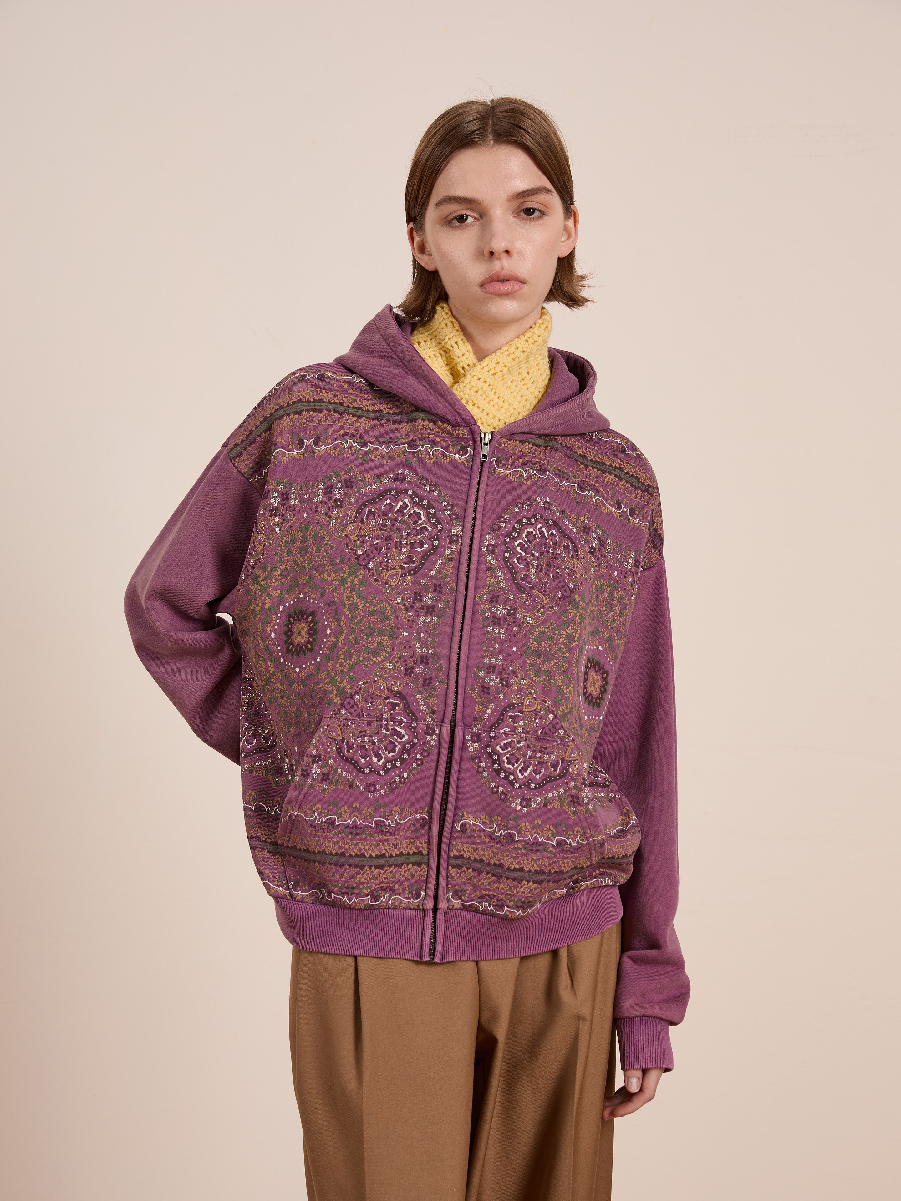 Against a beige background, an individual showcases the FOUND brand's Motif Zip-Up Hoodie with mandala-inspired purple patterns over a yellow sweater. The ensemble is completed with brown pants, offering a unique and stylish streetwear look.