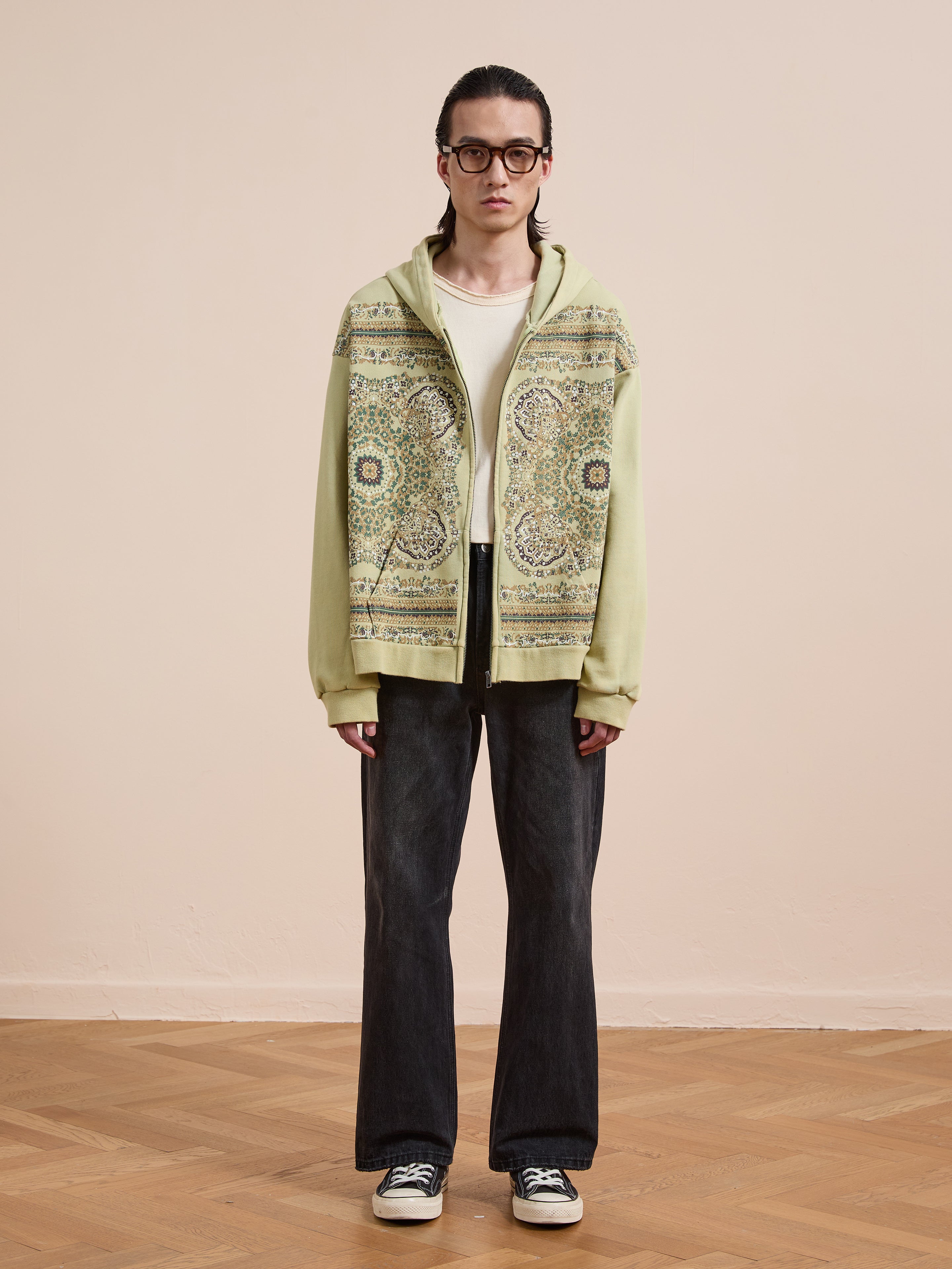 Person indoors wearing glasses and a FOUND Motif Zip-Up Hoodie in light green over a white shirt, paired with dark jeans and sneakers, standing against a beige wall and parquet floor.