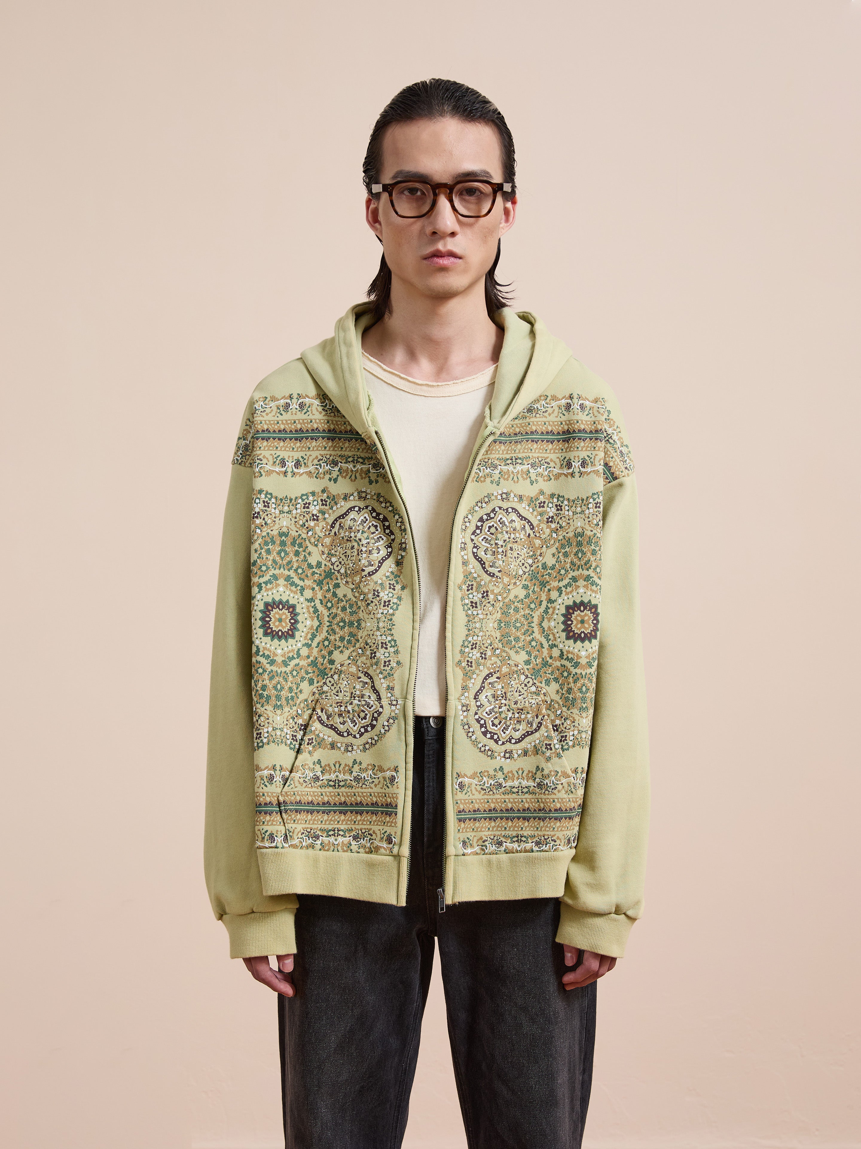A person in glasses wears the FOUND Motif Zip-Up Hoodie—a light, patterned streetwear essential—over an ivory shirt and dark pants, standing against a plain beige background.