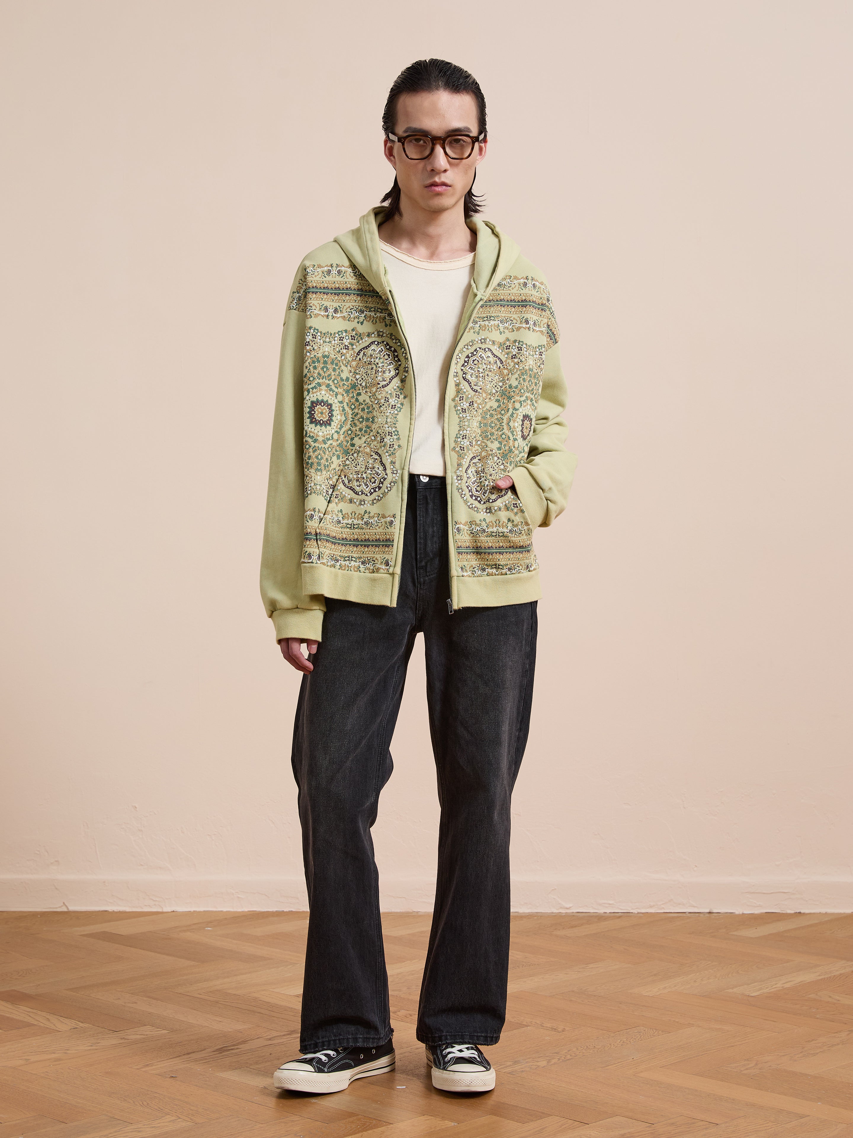 Indoors against a beige wall, a person wears glasses, a white shirt, dark wide-leg jeans, black sneakers, and the FOUND Motif Zip-Up Hoodie—a streetwear essential—on a wooden floor.
