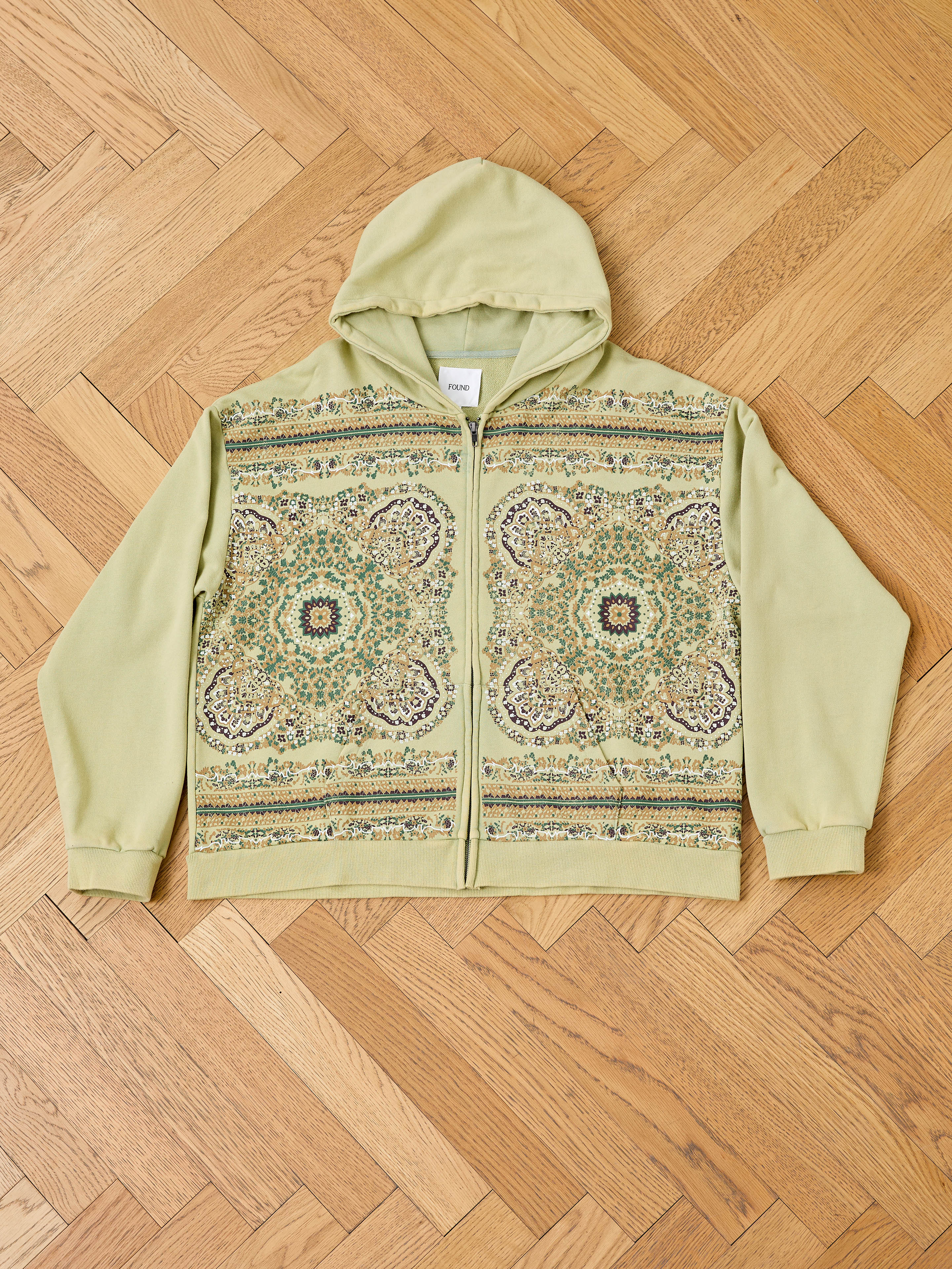 The FOUND Motif Zip-Up Hoodie, shown in light green on a wooden floor, features detailed floral and geometric patterns on the front—blending modern streetwear style with mandala art vibes.