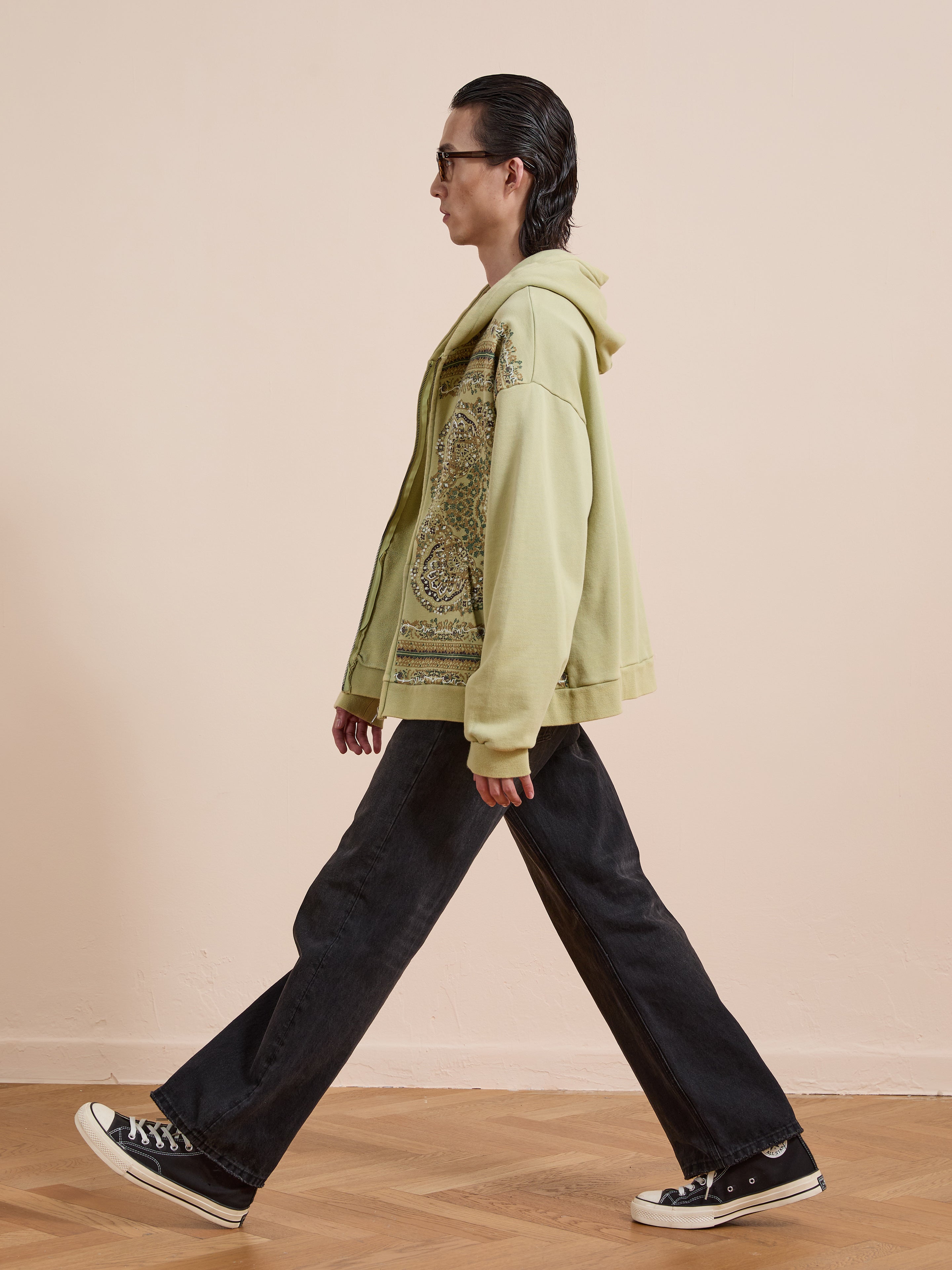 Wearing a light green FOUND Motif Zip-Up Hoodie, black flared jeans, and black sneakers, someone walks across a wooden floor by a beige wall—showcasing iconic streetwear style.