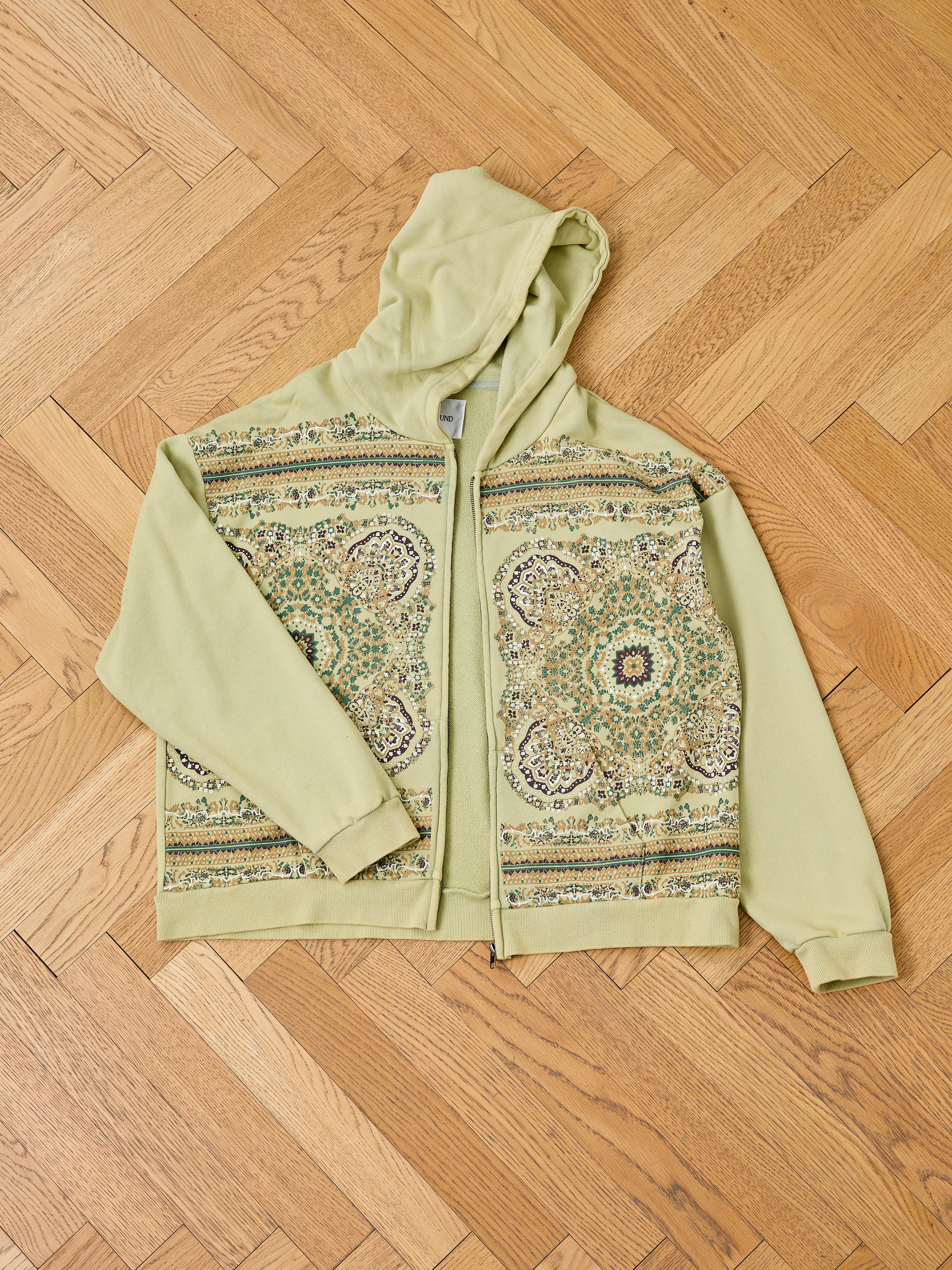 The FOUND Motif Zip-Up Hoodie in light green features an ornate, symmetrical floral and geometric design on the front—an essential streetwear piece, shown here laid out on a wooden parquet floor.