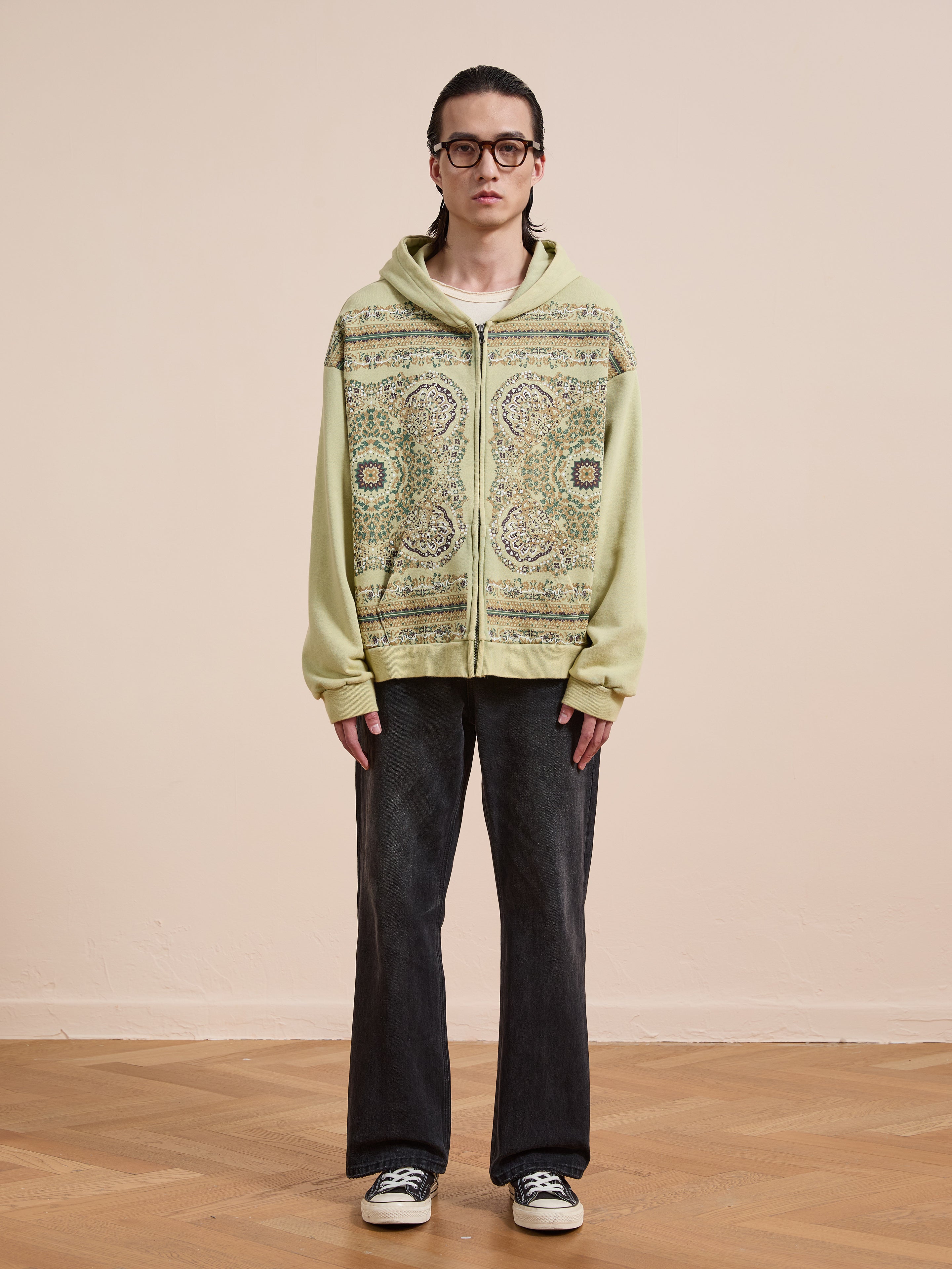 Person stands indoors against a plain beige wall, wearing glasses, the FOUND Motif Zip-Up Hoodie—a streetwear essential—dark wide-leg jeans, and black-and-white sneakers.