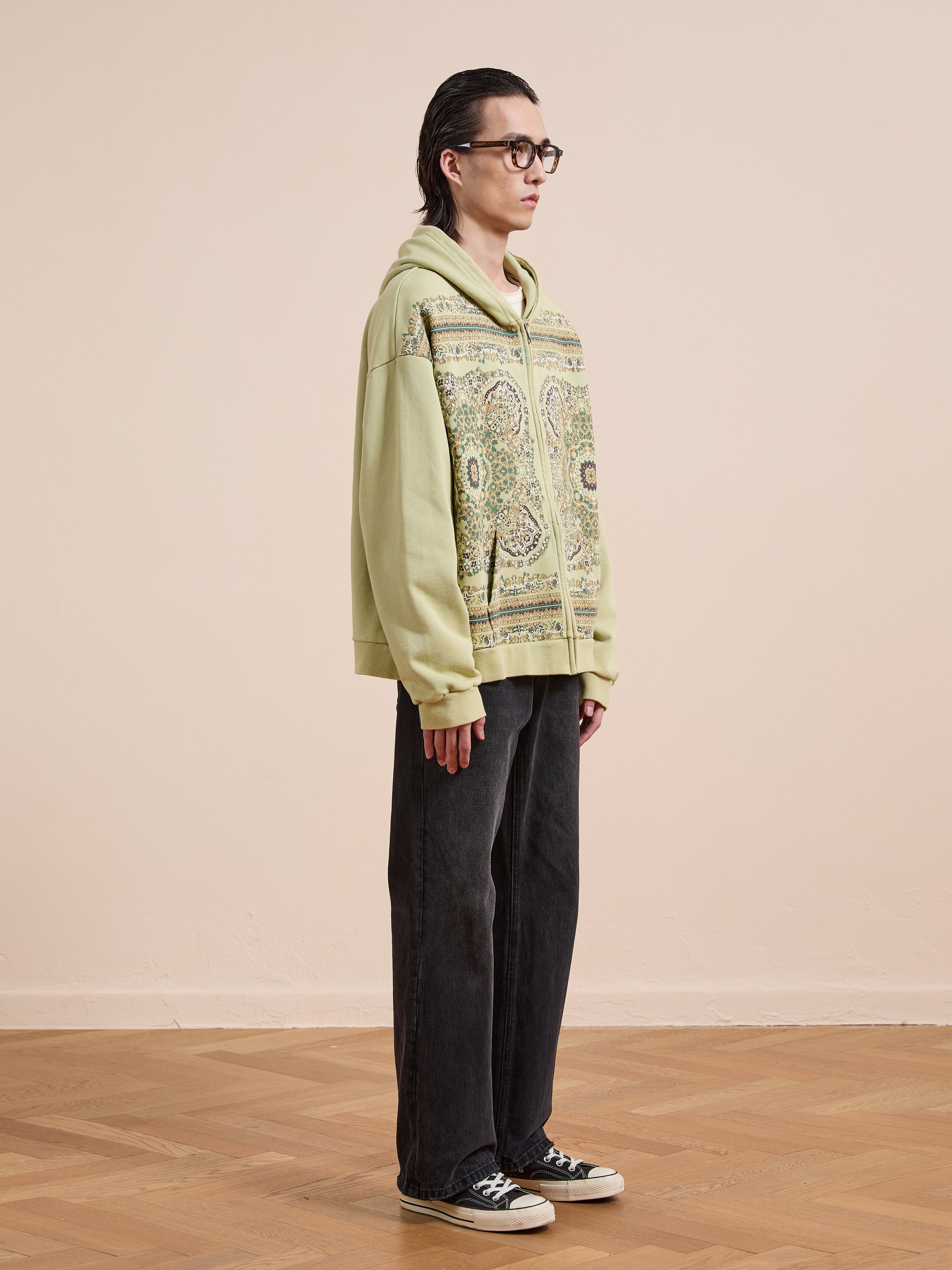 A person stands indoors on a wooden floor wearing glasses, black wide-leg pants, black sneakers, and the FOUND Motif Zip-Up Hoodie in light green, creating a stylish streetwear look.