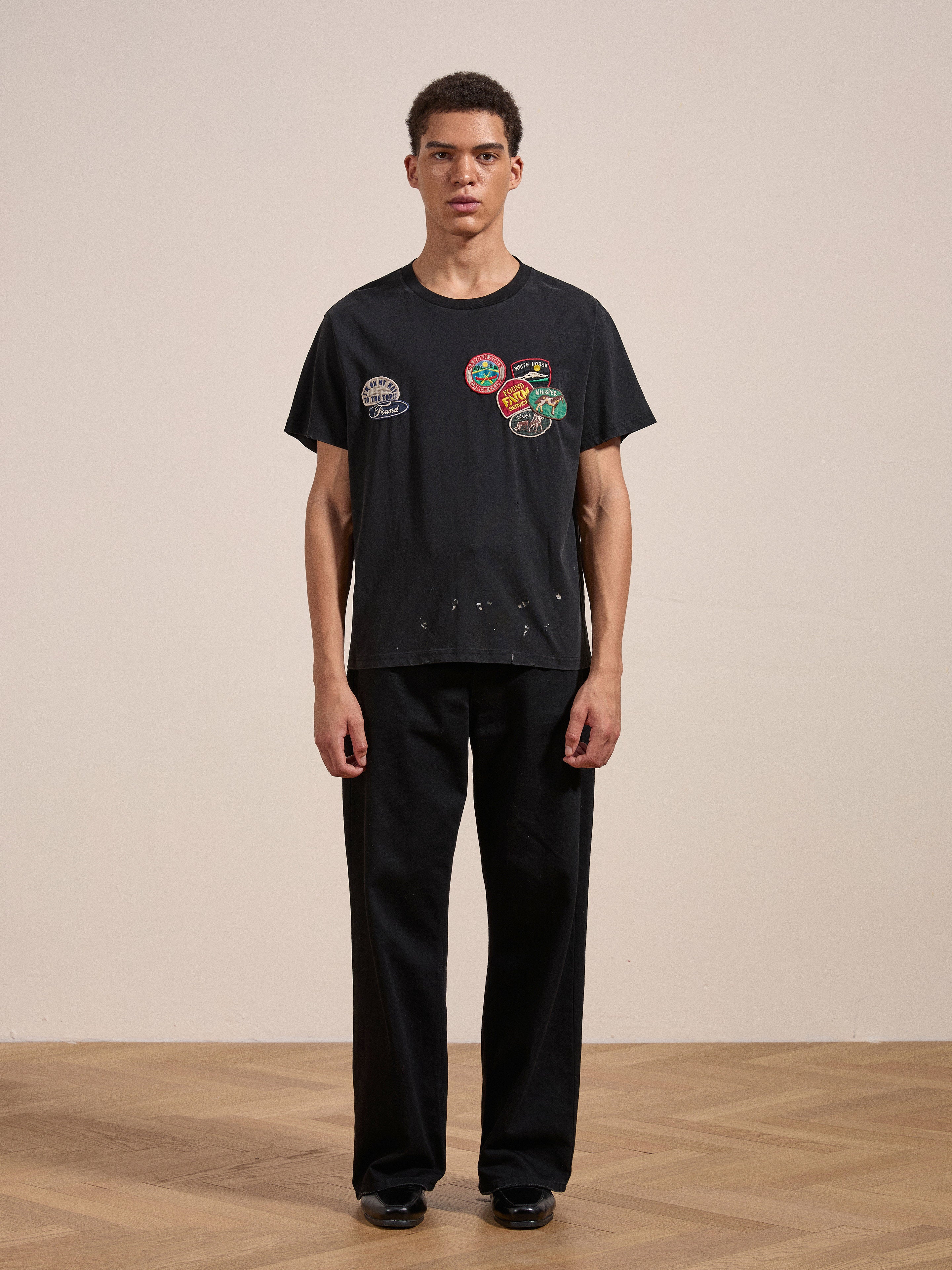 A person stands against a plain background, wearing the FOUND Multi Patch Washed Tee with various colorful patches on the front and black pants.