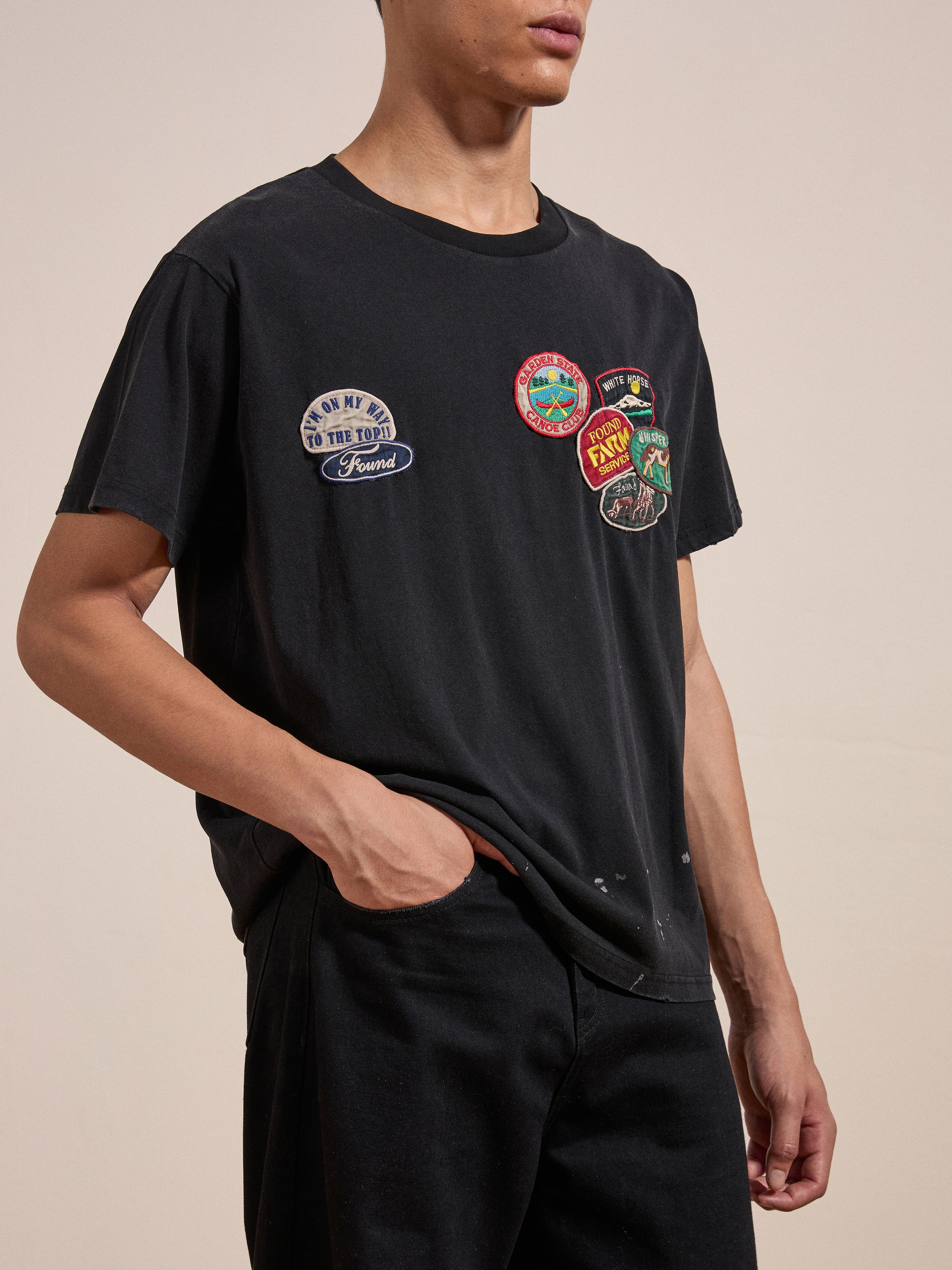 A person wearing the FOUND Multi Patch Washed Tee, featuring colorful patches on the left side, stands with one hand in their pocket against a neutral background.