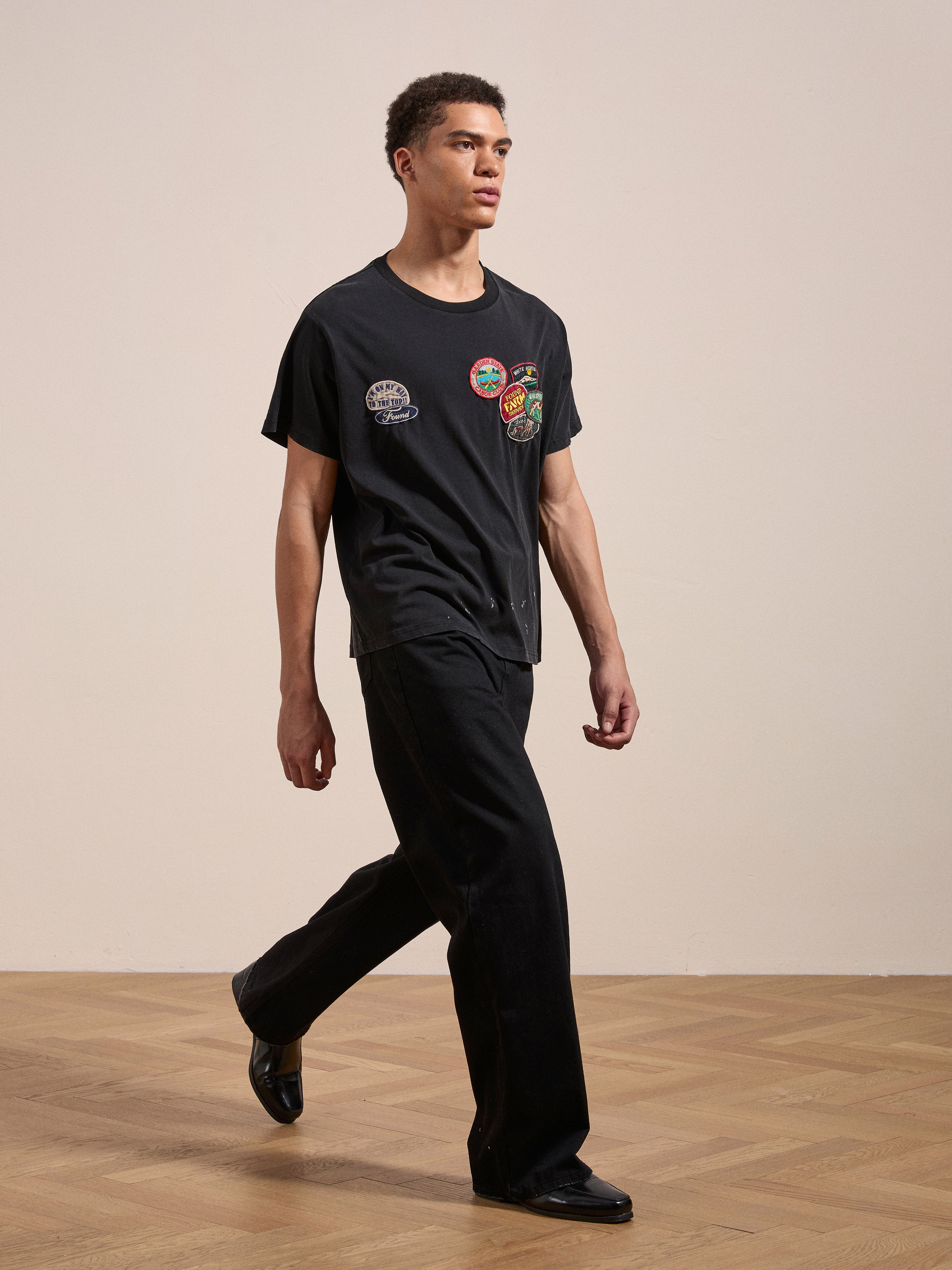 A man walks on a wooden floor wearing the FOUND Multi Patch Washed Tee and black pants. He has short hair and is facing slightly to the left.