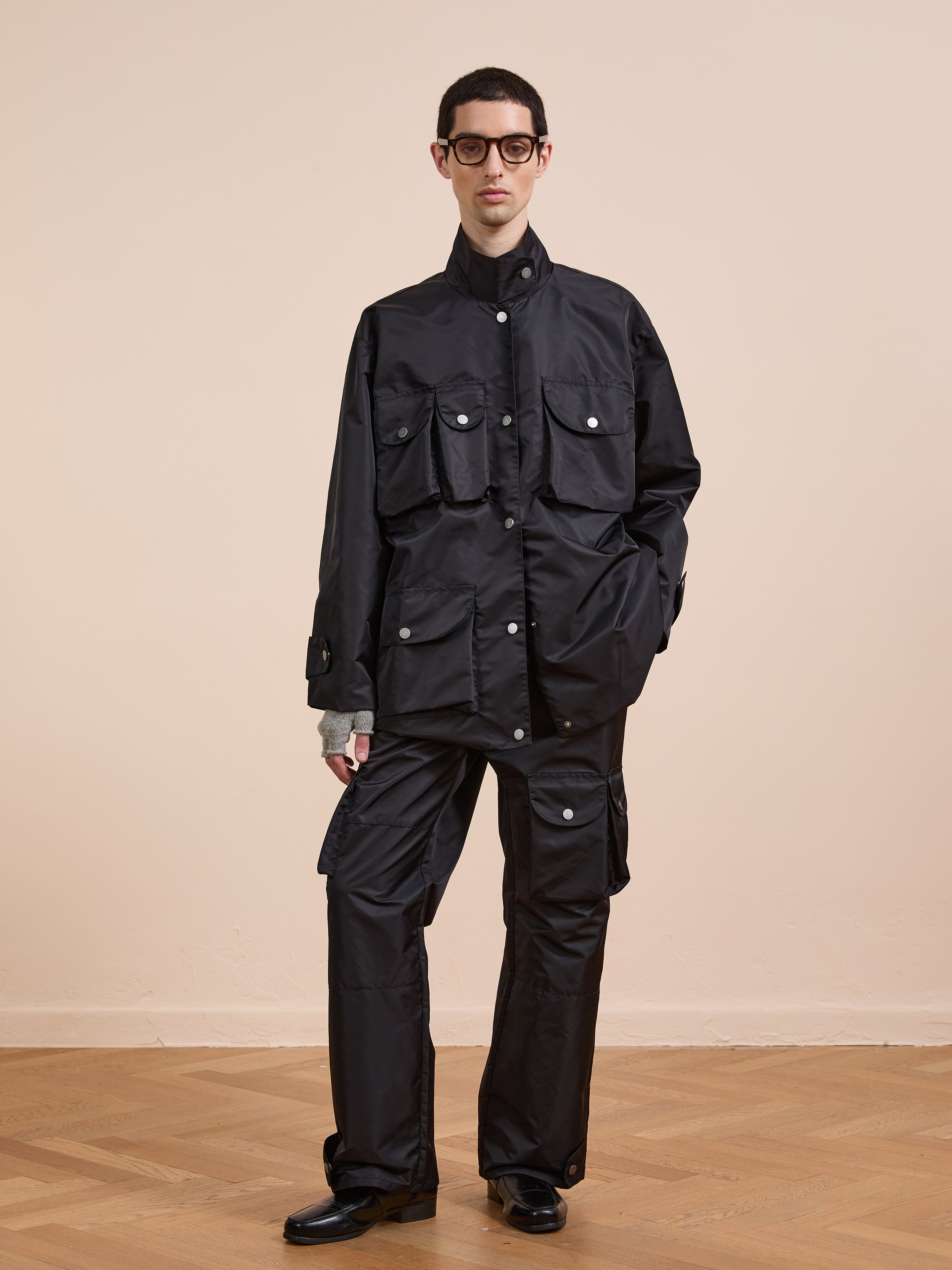 A person in the FOUND Cargo Nylon Jacket, paired with matching cargo pants and black shoes, stands on a wooden floor against a beige wall, wearing glasses and showing a neutral expression.