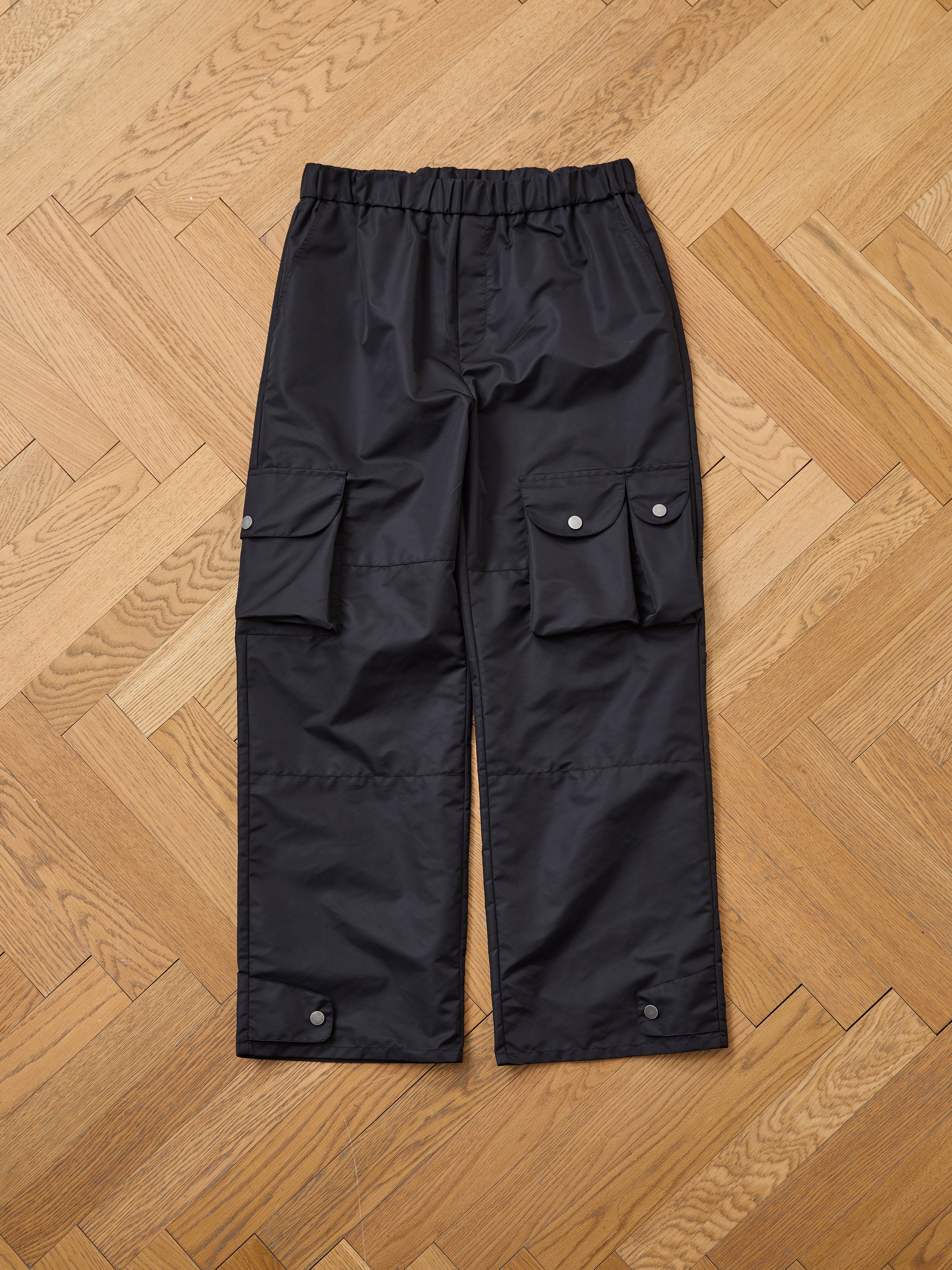 FOUND Nylon Cargo Pants in black, unisex with a relaxed fit, elastic waistband, and multiple flap pockets, displayed flat on a wooden herringbone floor.