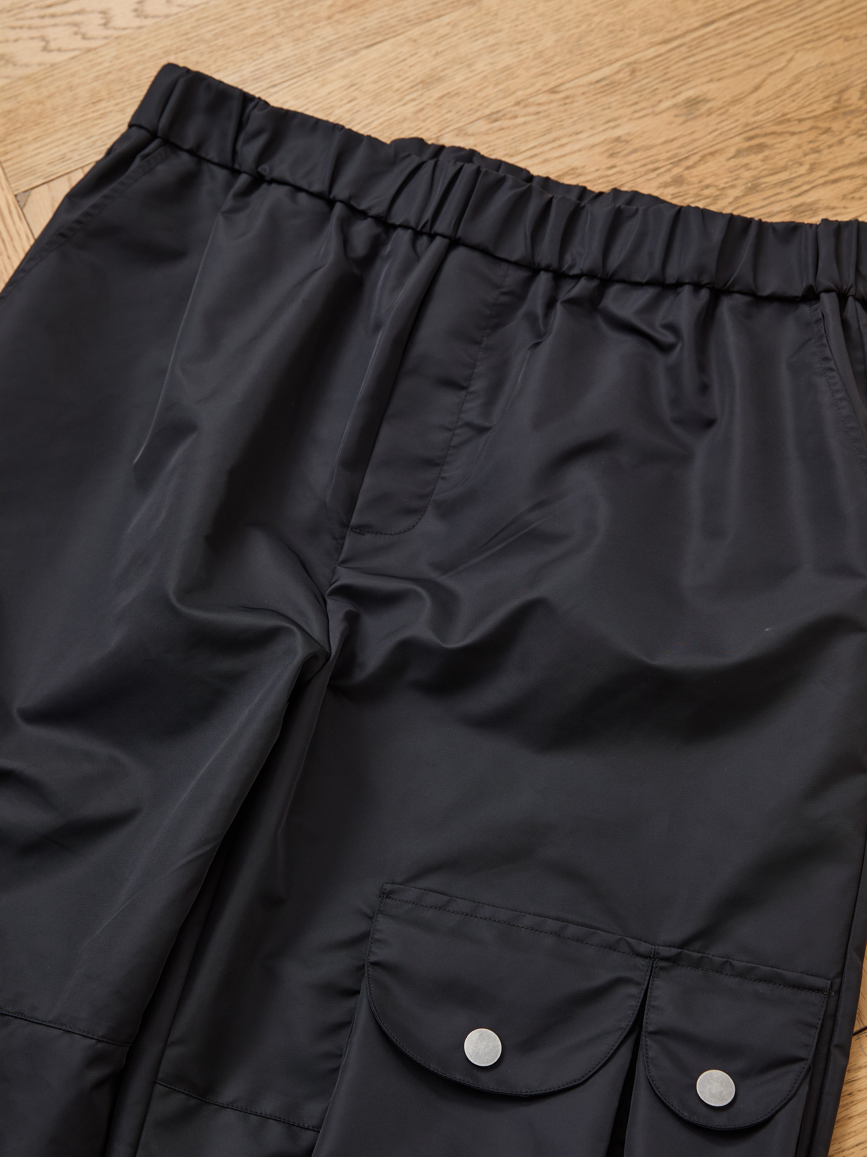 FOUND Nylon Cargo Pants in black, unisex design, relaxed fit with an elastic waistband and flap pockets with snap buttons, shown laid on a wooden floor.