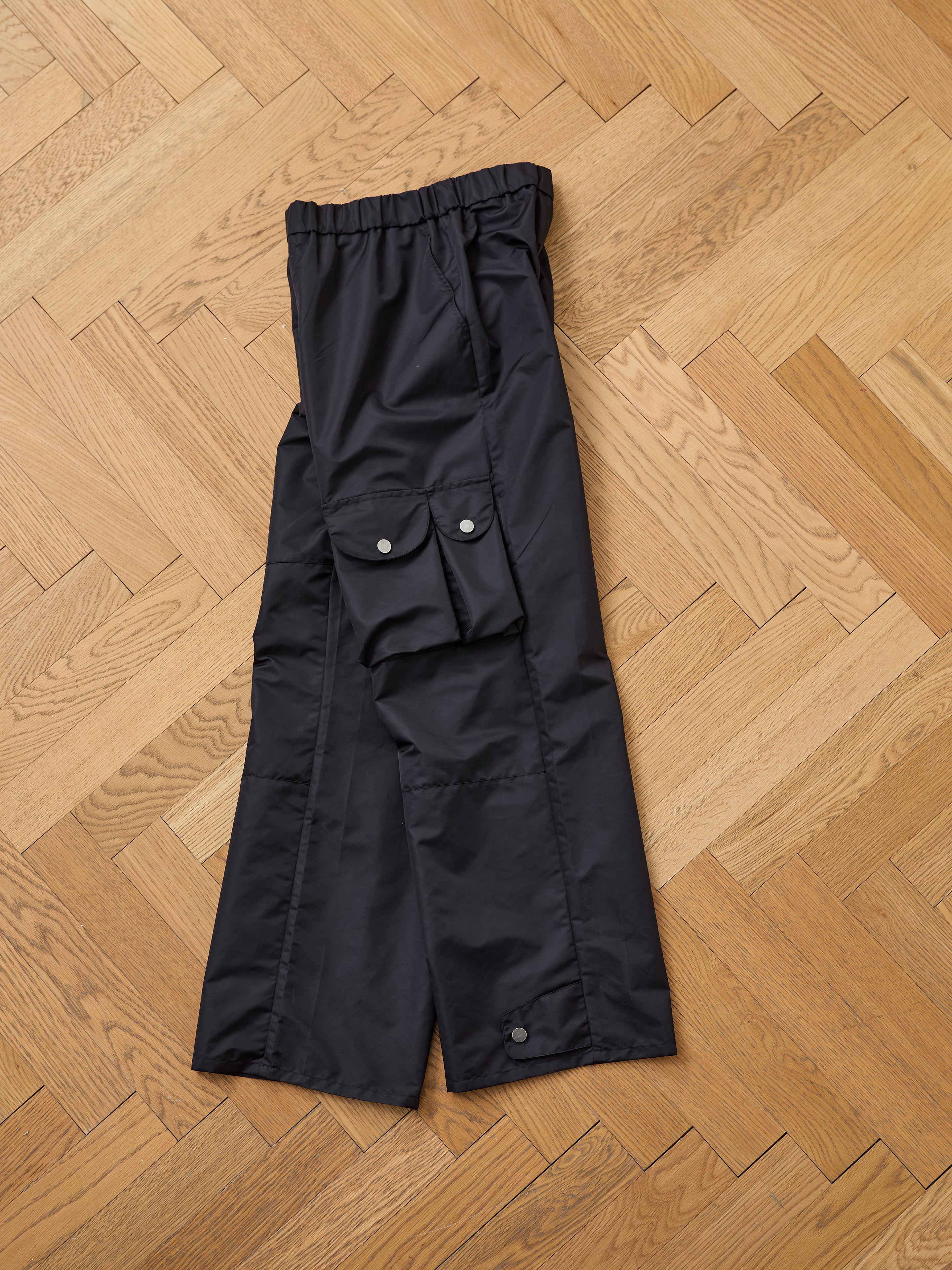 FOUND Nylon Cargo Pants — unisex, black, relaxed fit with flap pockets and snap buttons, made from nylon. Shown laid flat on a light-colored wooden herringbone floor.