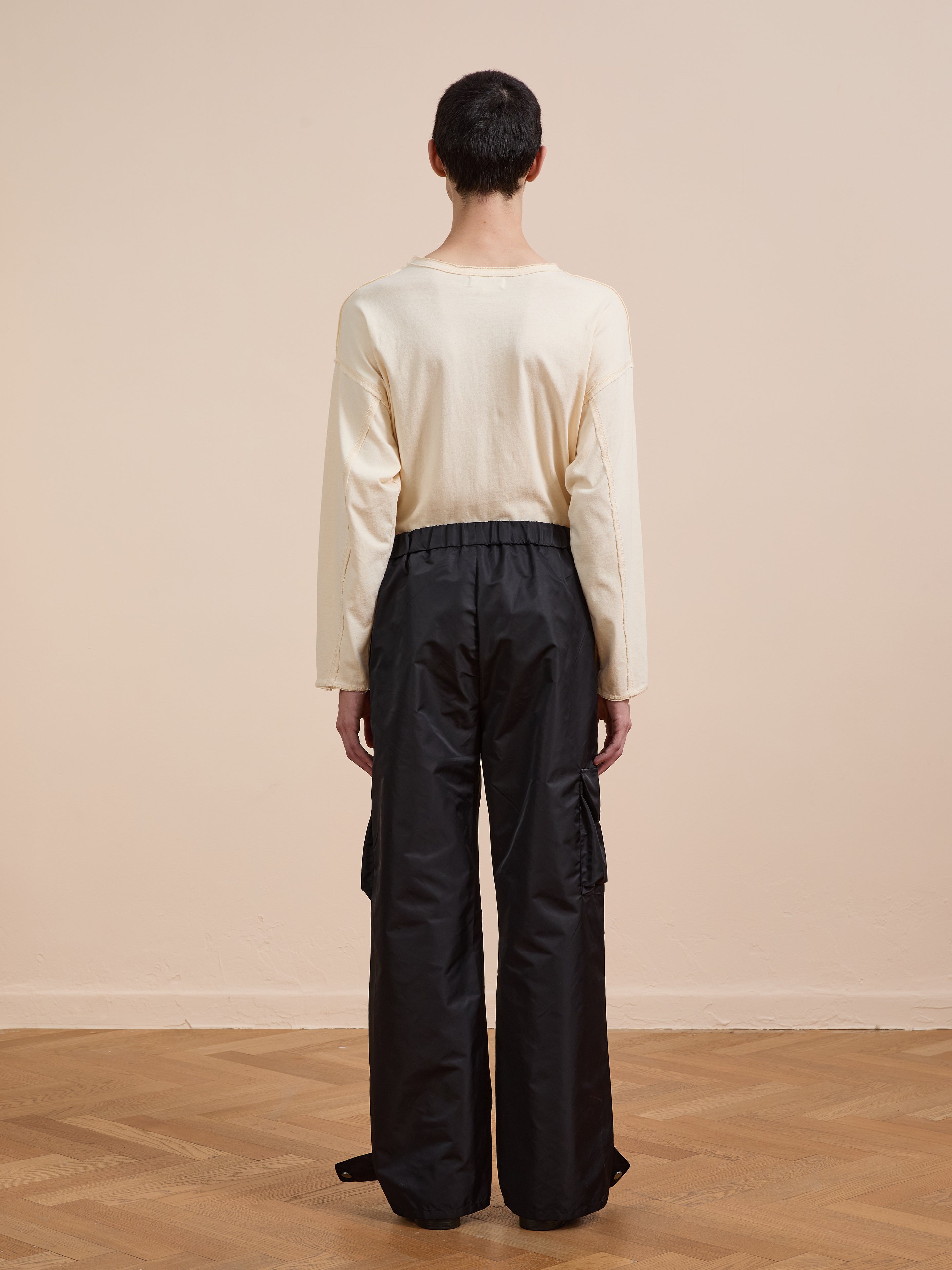A person with short dark hair, in a long-sleeve cream shirt and FOUND Nylon Cargo Pants, stands facing away on a wooden floor against a plain beige wall.