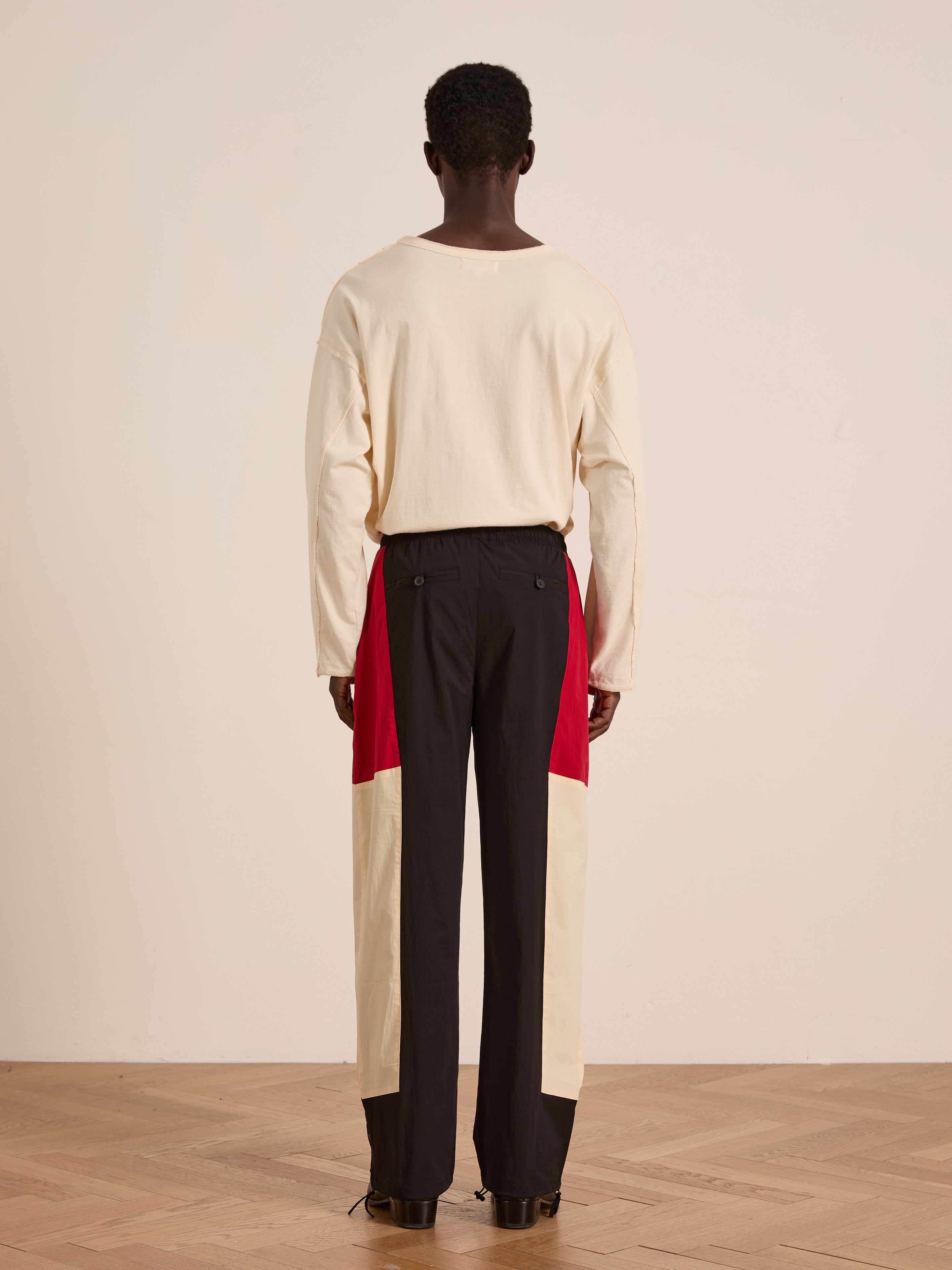 A person stands facing away, wearing a long-sleeve cream shirt and Found’s Pine and Rose Track Pants—unisex, relaxed fit nylon pants with red and cream color block panels—on a wooden floor against a plain wall.