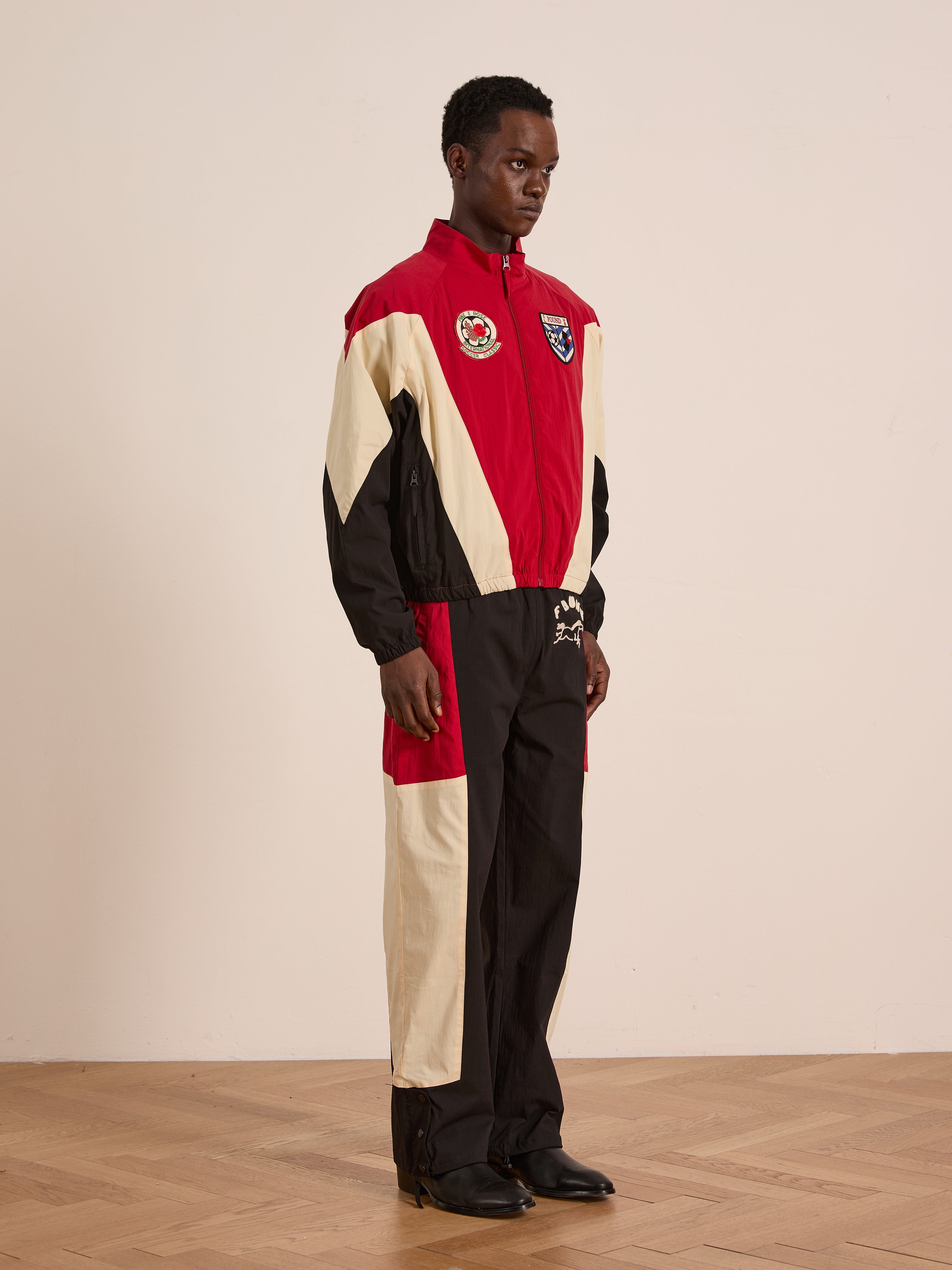 Someone stands indoors on a wooden floor against a plain light wall, wearing Found’s Pine and Rose Track Pants—unisex relaxed-fit with red, black, and beige color-blocking and embroidered patches.
