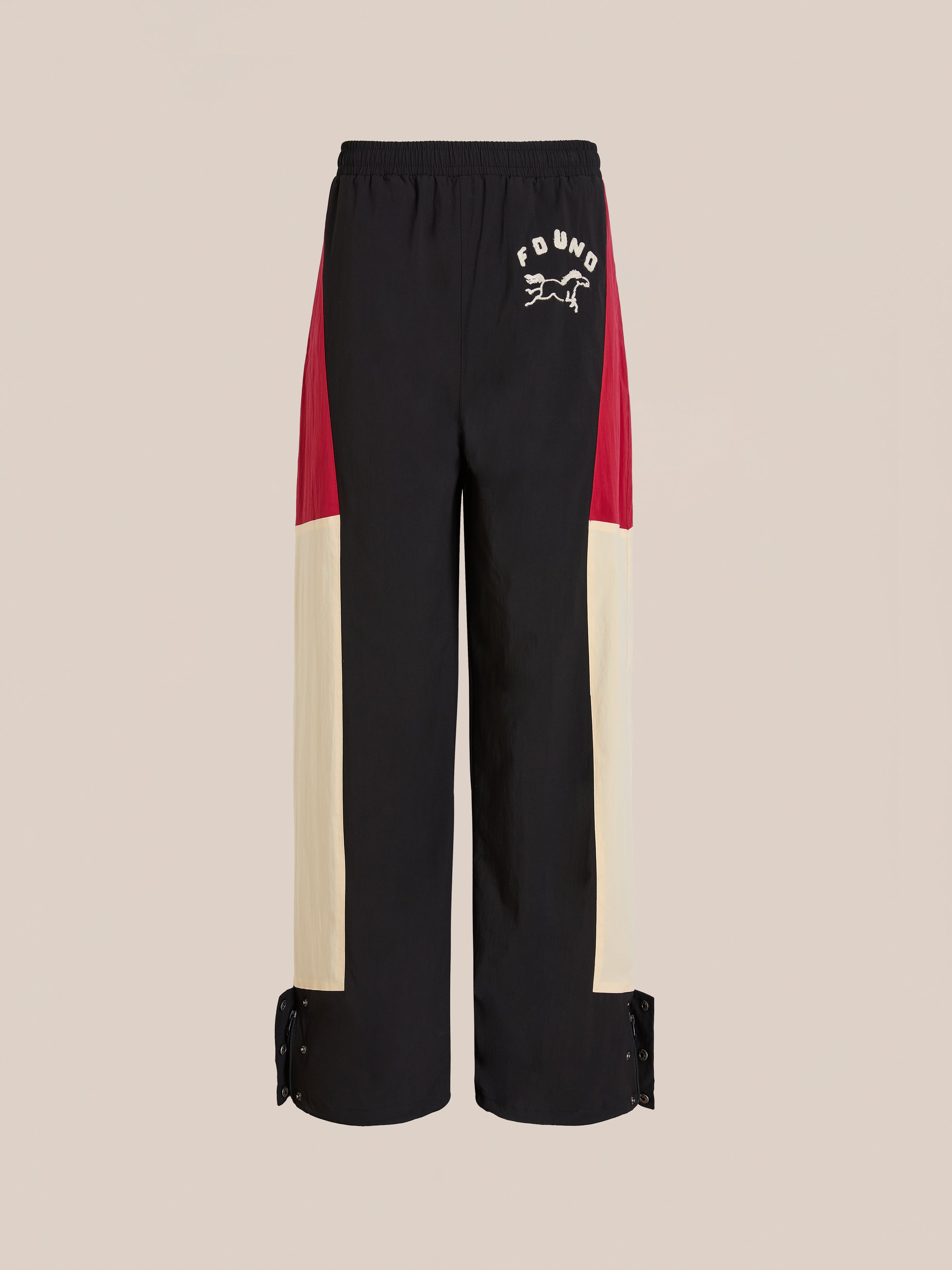 Found Pine and Rose Track Pants feature cream and red panels, elastic waistband, snap button cuffs, unisex relaxed fit, and "FOUND" with a running animal graphic on the upper left thigh.