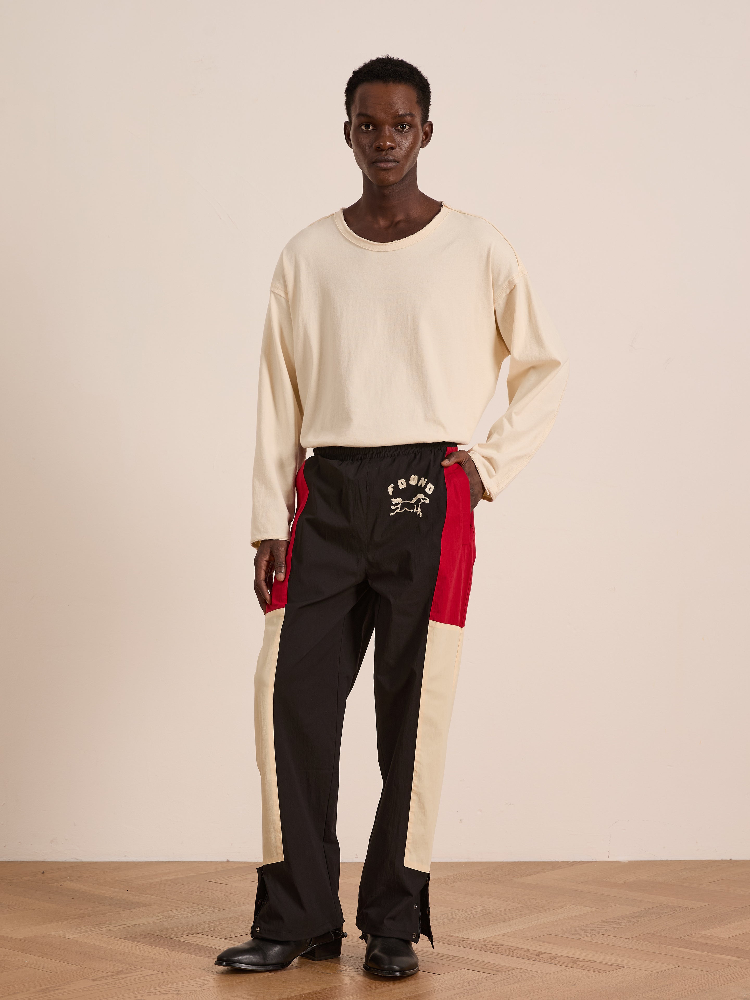 A person stands indoors wearing a long-sleeve cream shirt, Found Pine and Rose Track Pants with cream and red panels in a relaxed unisex fit, and black shoes against a plain background.