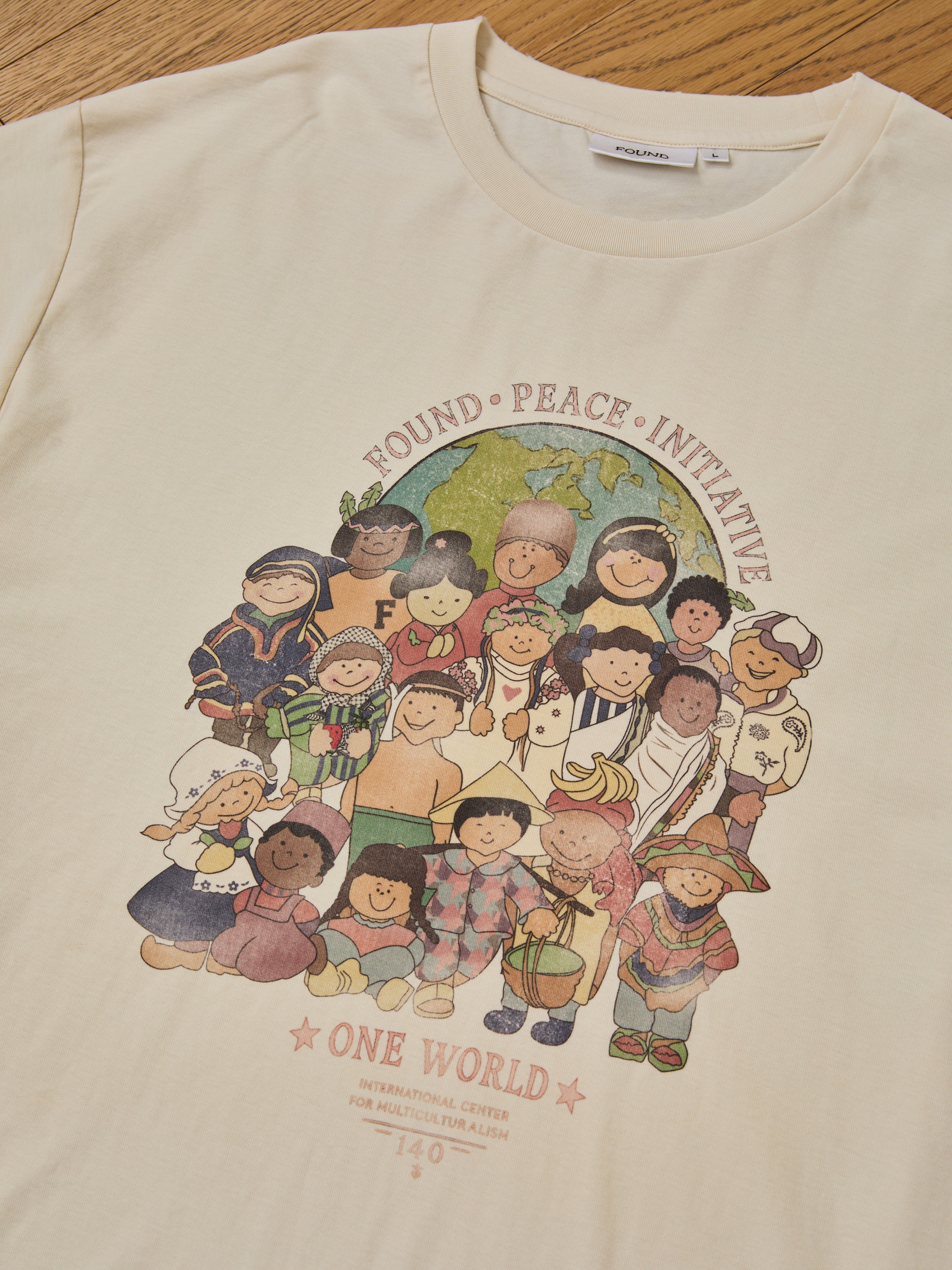 The cream One World Family Tee by FOUND features diverse children in cultural attire and "FOUND PEACE INITIATIVE" and "ONE WORLD" prints, celebrating unity and multiculturalism.
