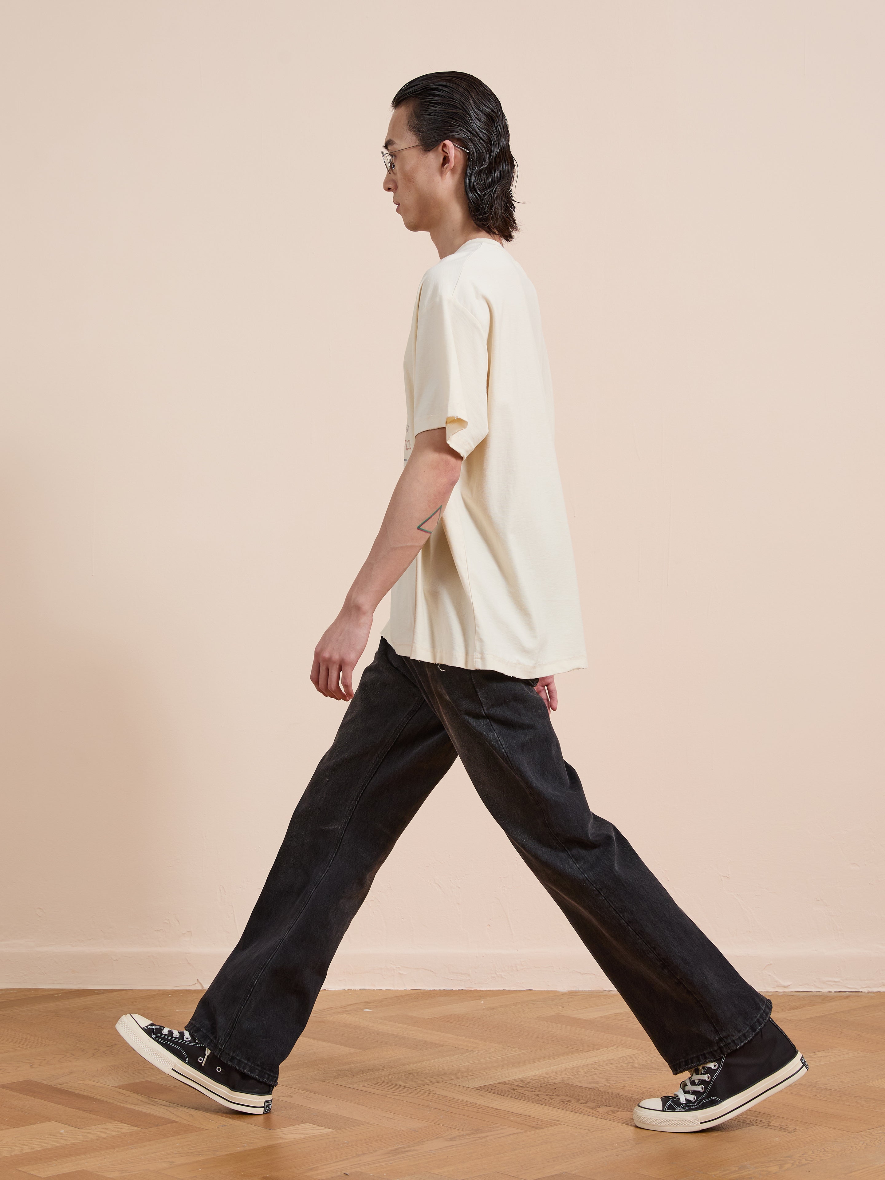 A person with long dark hair, wearing FOUND’s white One Humanity Tee and black jeans, walks in profile on a wooden floor against a beige wall.