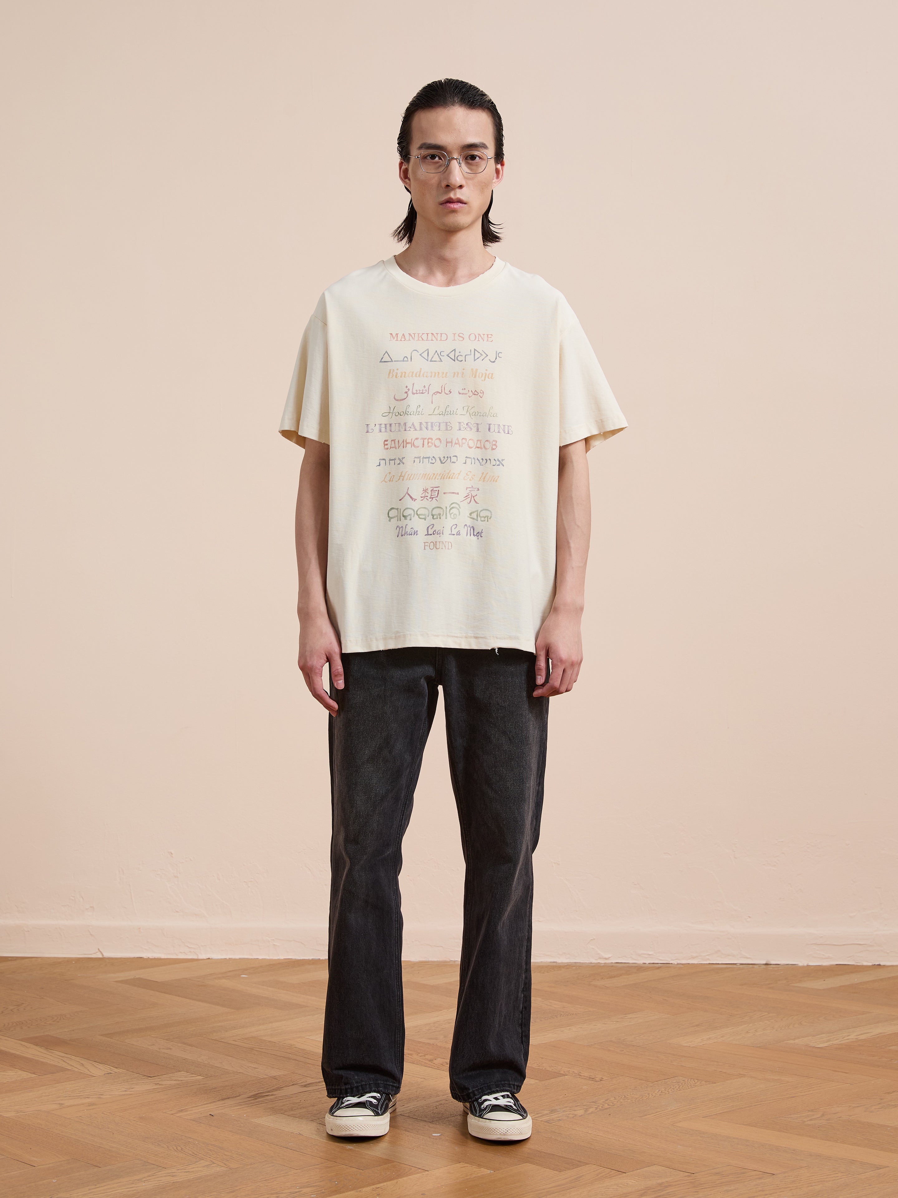 A person with long hair and glasses stands facing forward on a wooden floor against a beige wall, wearing the FOUND One Humanity Tee—a vintage cotton unisex relaxed fit graphic t-shirt—dark jeans, and black sneakers.