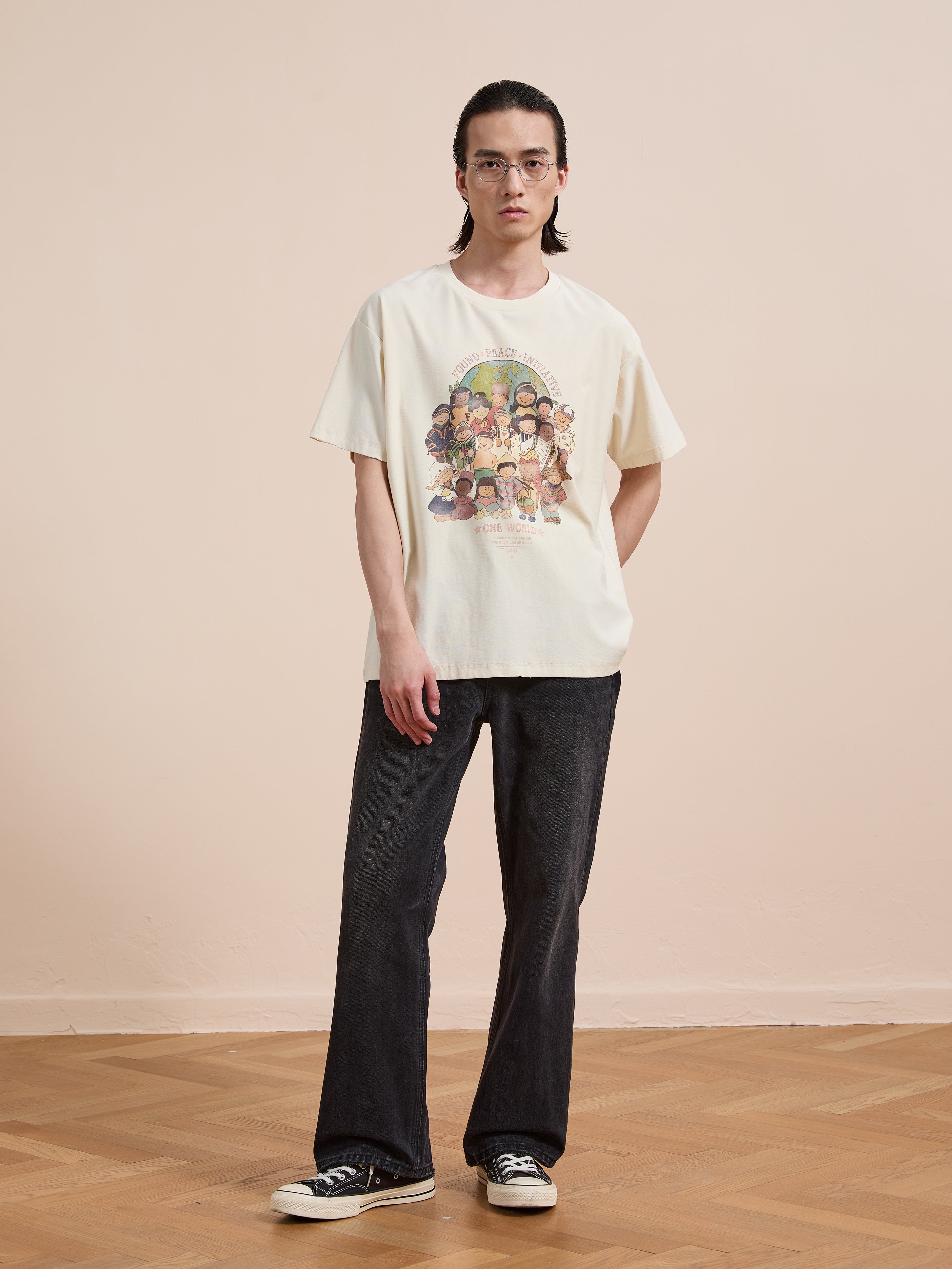 A person with long dark hair, glasses, dark jeans, and black sneakers wears the FOUND One World Family Tee while standing on a wooden floor against a plain beige wall.
