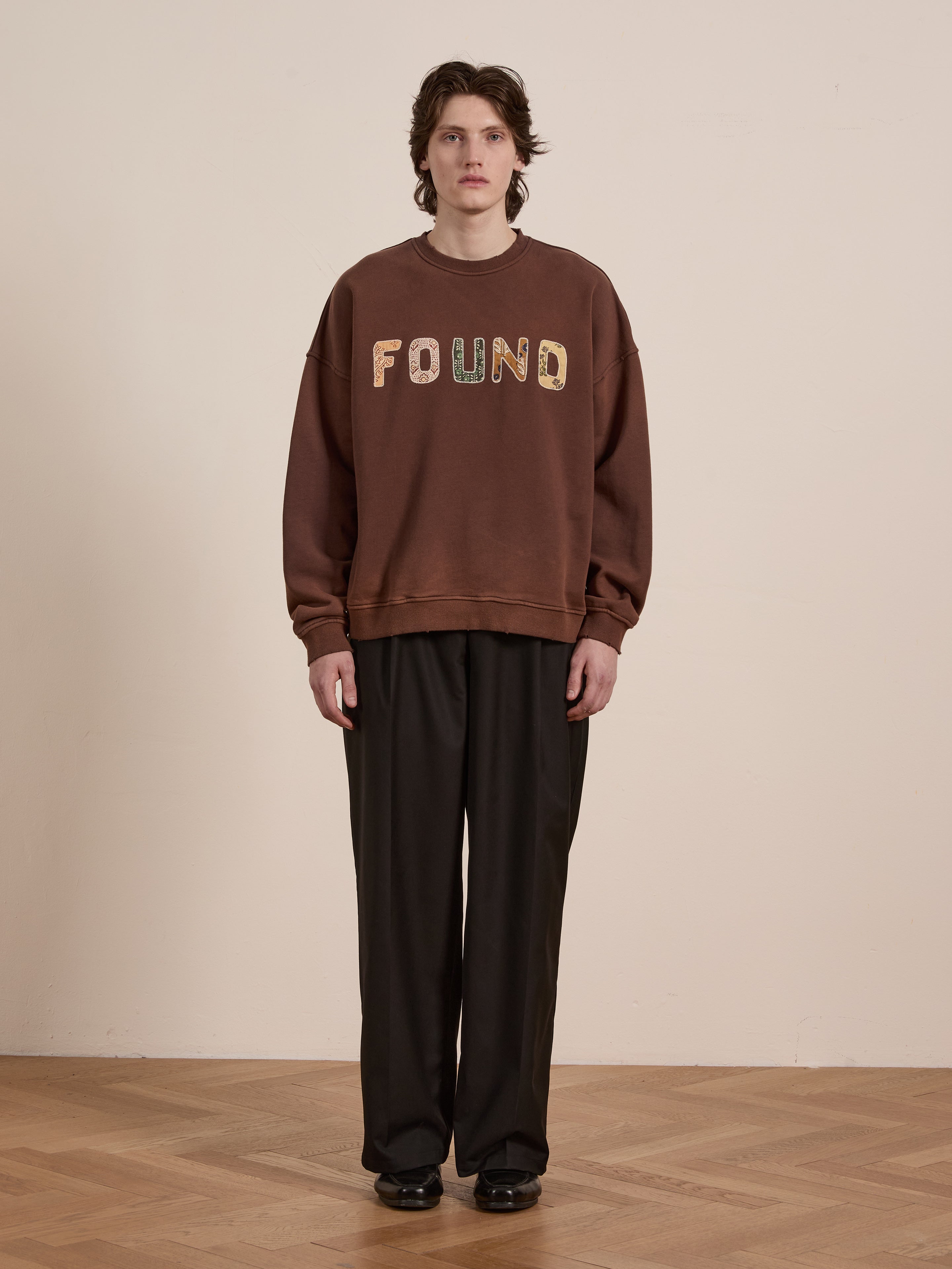 A person stands on a wooden floor wearing the FOUND Patterned Logo Crewneck with "FOUND" appliqué lettering, made from soft French terry cotton, paired with dark pants.