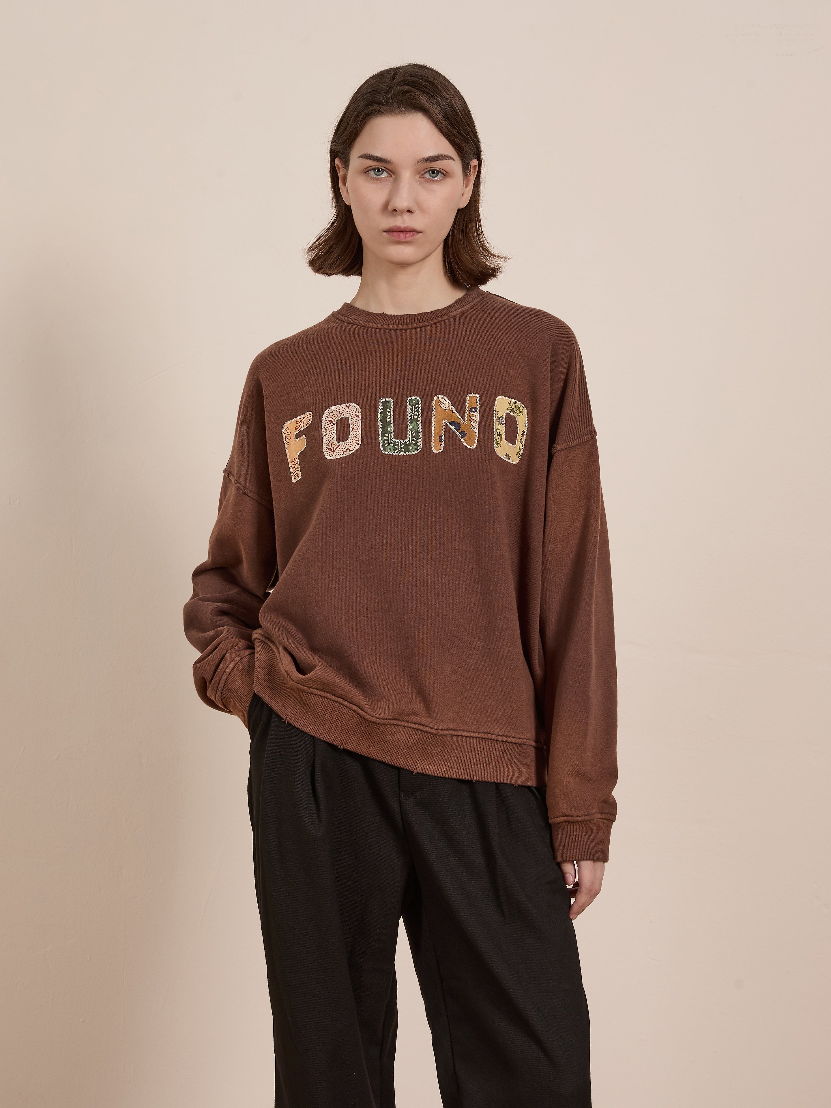 A person is wearing a FOUND Patterned Logo Crewneck with "FOUND" appliqué lettering against a plain background, paired with black trousers.