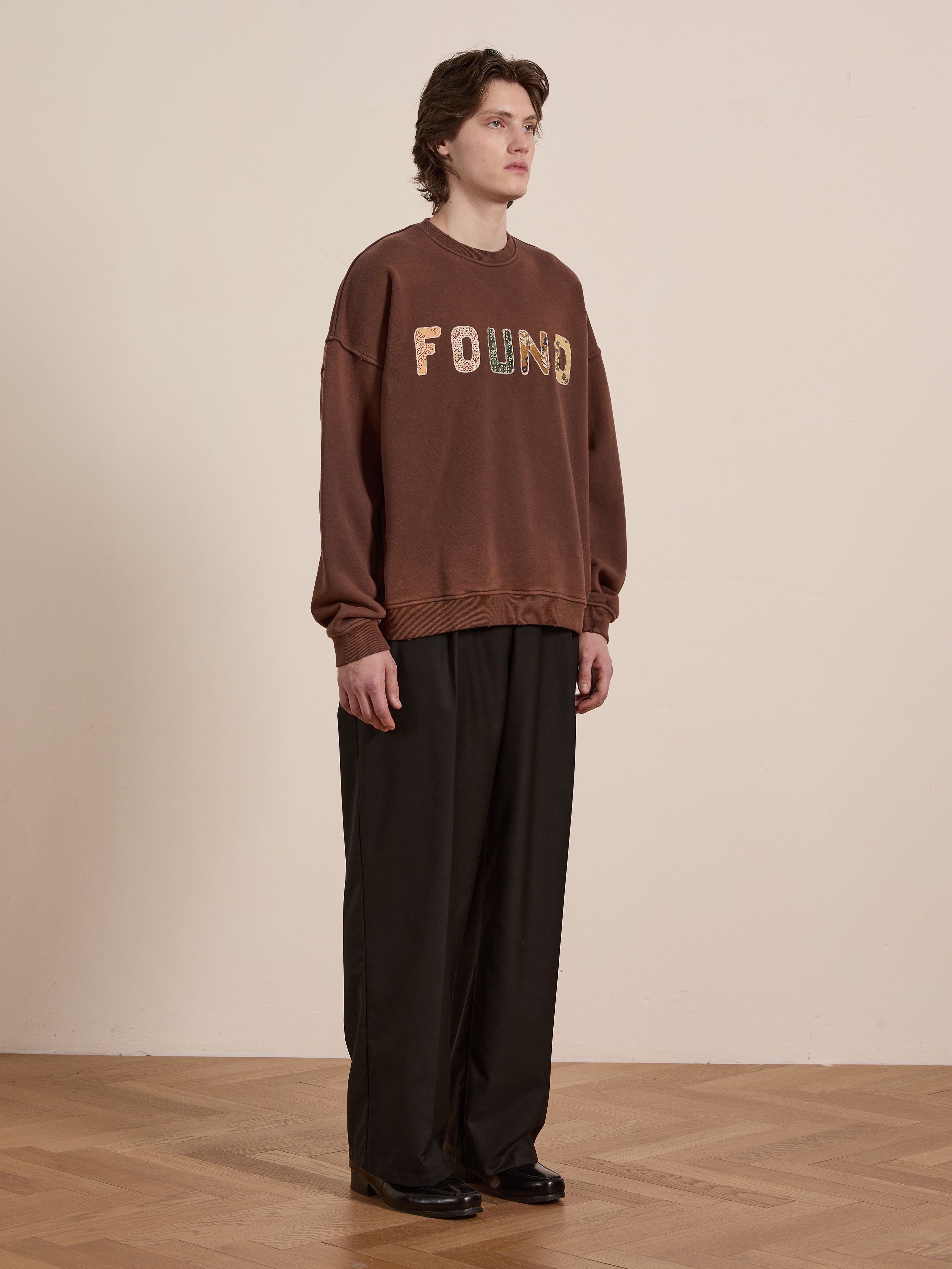 A medium-haired person stands against a plain backdrop, wearing a FOUND Patterned Logo Crewneck made from French terry cotton with "FOUND" appliqué lettering and loose dark trousers, looking to the side.