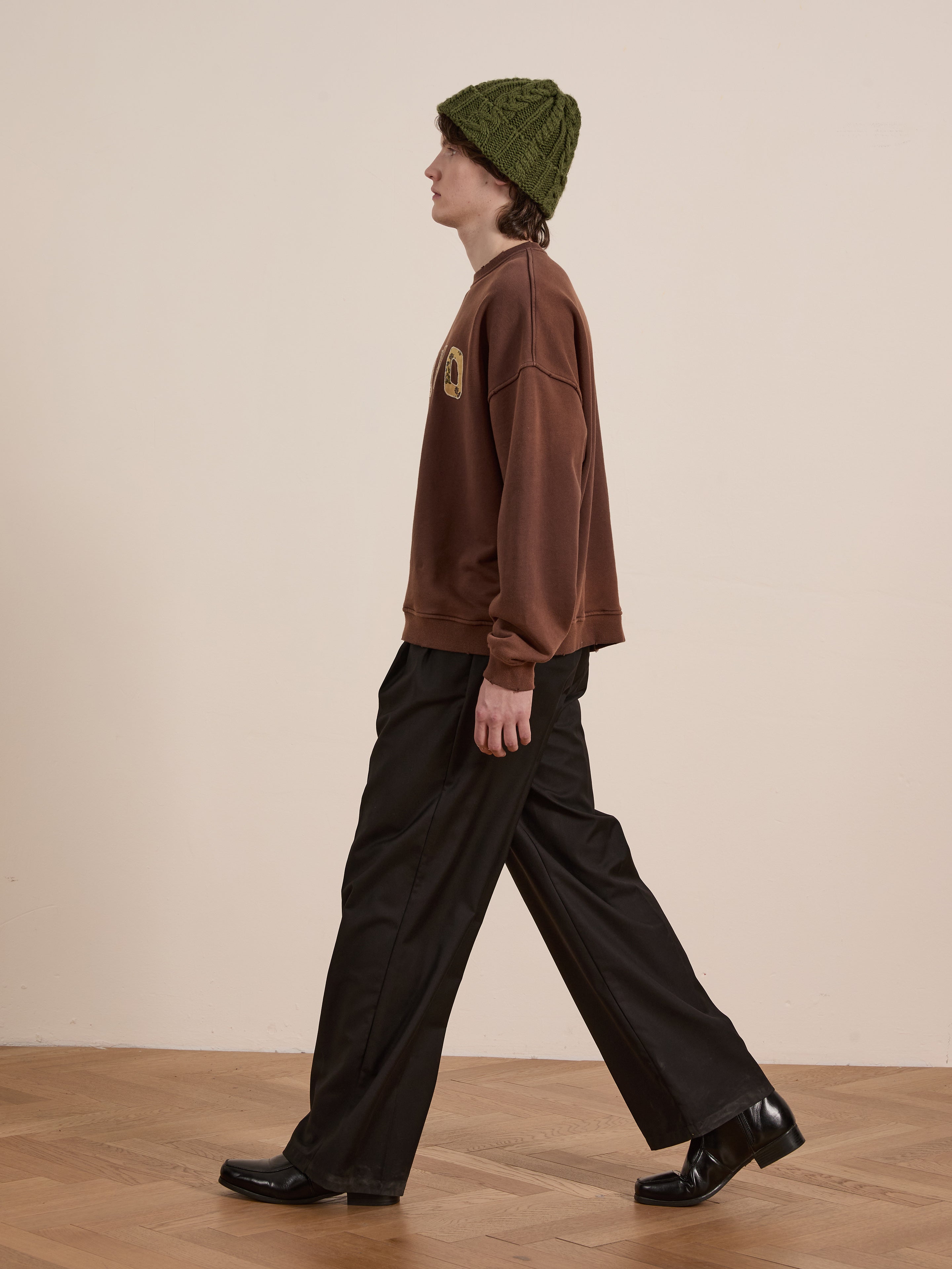 A person in a FOUND Patterned Logo Crewneck made from French terry cotton, black pants, black shoes, and a green knit hat is walking sideways on a wooden floor.